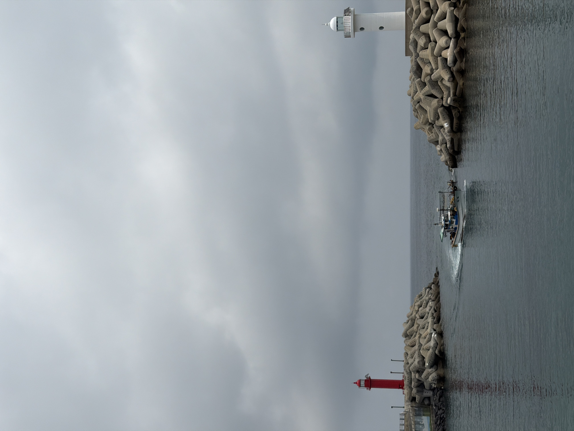 A fishing boat with a white hull and green superstructure is moving towards the viewer from between two breakwaters in the sea. The breakwaters are constructed of large, gray, concrete tetrapods. On the left, a red lighthouse stands tall on a pier, and on the right, a white lighthouse is visible on another breakwater. The sky is overcast and gray. This scene appears to be the entrance to a harbor in Jeju, South Korea.