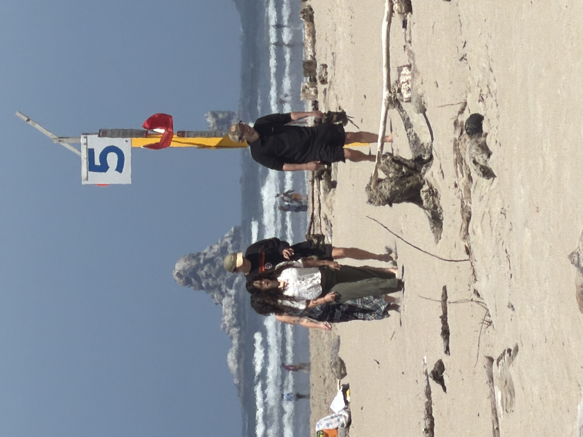 In Cannon Beach, Oregon, a group of four people are walking on a sandy beach. In the background, there are large rock formations jutting out of the ocean, with waves breaking. The sky is clear and blue. One person is wearing a black t-shirt and shorts, another is wearing a black long-sleeved shirt and shorts, and two women are wearing patterned dresses and green pants respectively. A yellow pole with a white sign displaying the number "5" and a red object hanging from it is visible in the foreground. There are also several pieces of driftwood scattered across the sand.