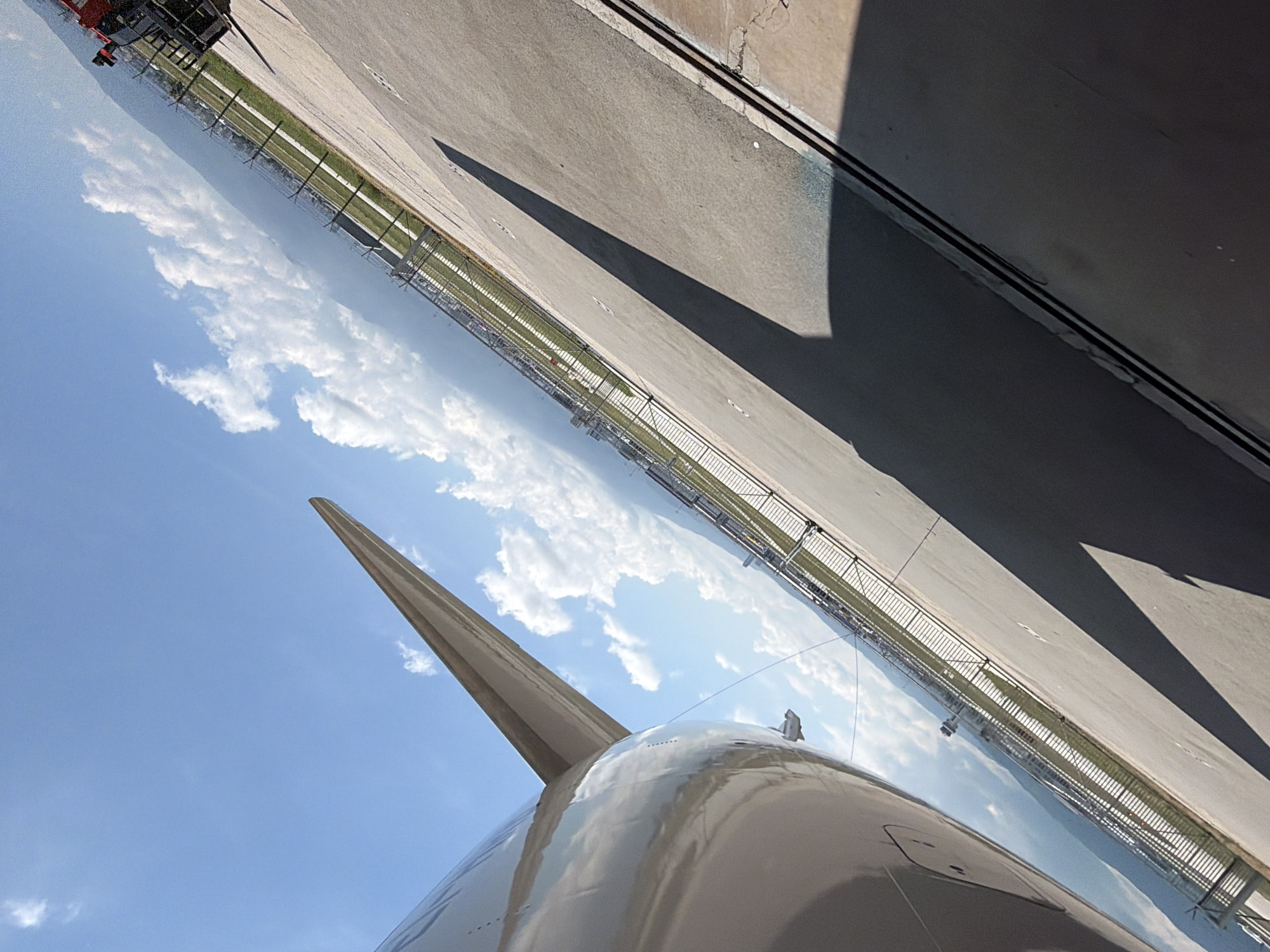 This image, taken in Sofia, Bulgaria, captures a view of a large aircraft parked on an airport tarmac under bright daylight. A significant portion of the aircraft's body, possibly an engine nacelle or part of the fuselage, dominates the lower and left-center sections of the frame, appearing reflective and light-colored. A sleek, pointed structure, resembling the leading edge of an engine intake or a winglet, extends upwards from the main aircraft body into the sky.
The sky, visible in the upper-left and central portions of the image, is a clear blue with scattered, fluffy white cumulus clouds. To the right, the grey asphalt or concrete tarmac of the airport is prominent, with long, dark shadows cast across its surface, indicative of strong sunlight. A tall metal fence, possibly chain-link or mesh, stretches horizontally across the middle ground, separating the tarmac from a narrow strip of green grass and distant, indistinct structures.
The aircraft appears stationary on the apron or a taxiway. No people or discernible activity are visible. The bright illumination and sharp shadows suggest a sunny mid-day. There is no visible text in the image.