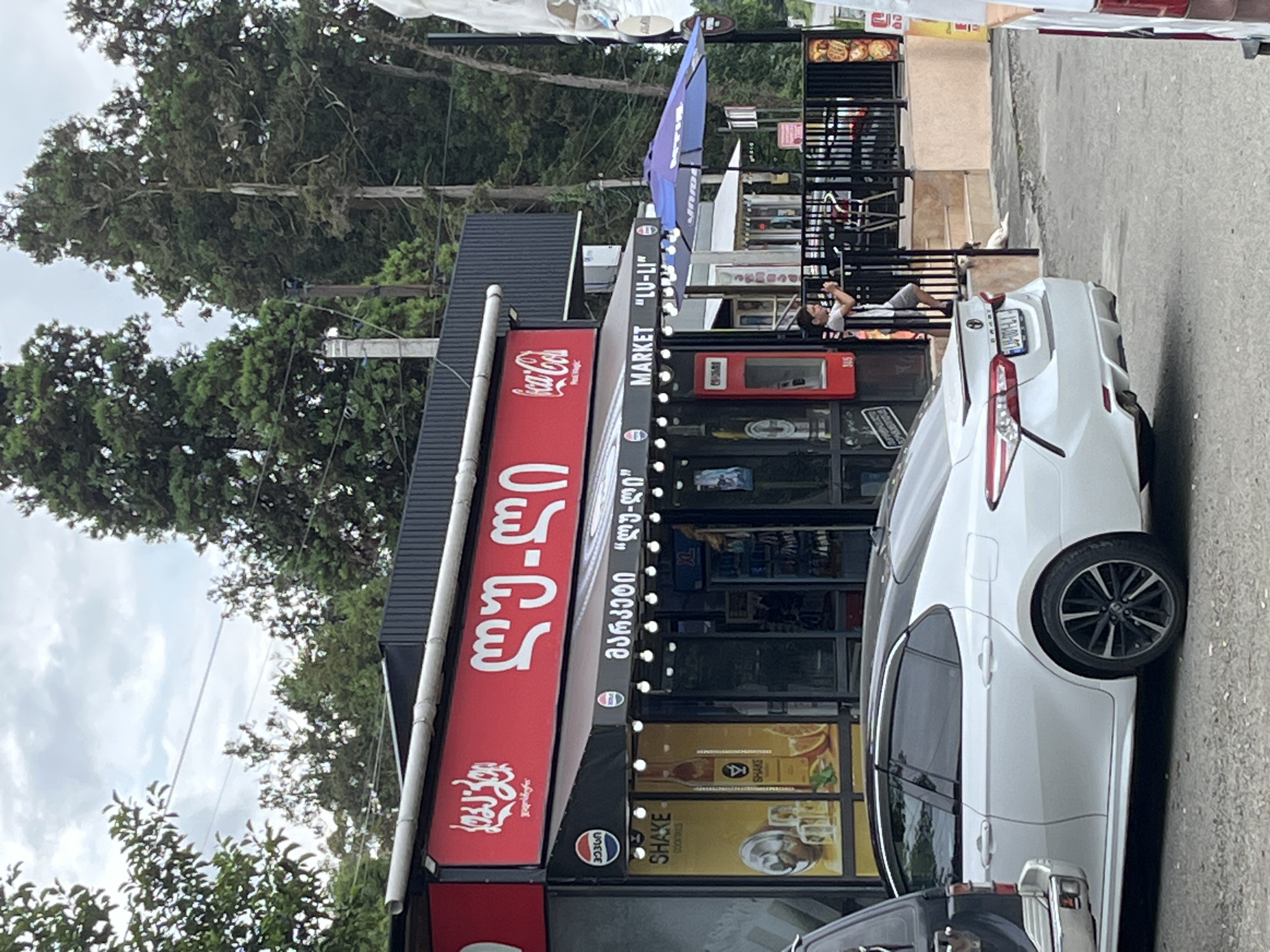 The image depicts a street-level view of a small market and cafe located in Ureki, Georgia. The building features a prominent red awning with white Georgian text and the Coca-Cola logo. Below the awning, a dark-colored canopy displays "MARKET 'LU-LI'" in white lettering, along with Pepsi logos. The storefront has large glass windows showcasing various products, including what appears to be drinks and snacks, with a "SHAKE COCKTAILS" advertisement. Outside the market, there's a black metal fence and a blue umbrella. A young person in a light-colored t-shirt and shorts is standing near the fence, holding a phone. In the foreground, a white sedan is parked. The background consists of tall, dark green evergreen trees and a cloudy sky.