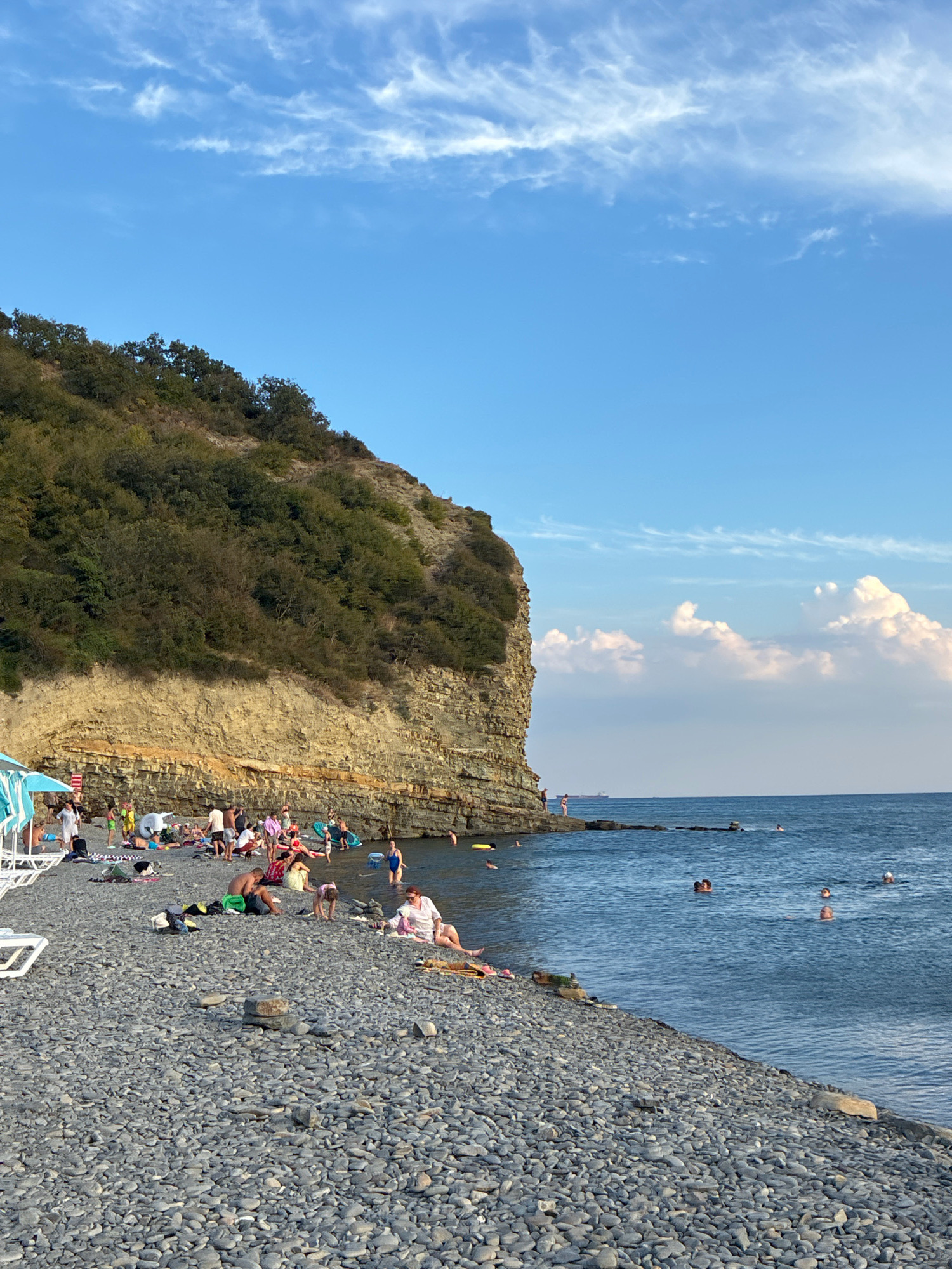In Dyurso, Russia, a crowded pebble beach is situated at the base of a steep, layered cliff face covered in dense green vegetation. The sea, a deep blue, stretches towards the horizon under a bright sky with scattered white clouds. Many people are visible on the beach, some relaxing on lounge chairs under blue and white umbrellas, while others are gathered in groups. Several individuals are in the water, swimming and enjoying the sea. The terrain of the beach is composed of smooth, grey pebbles. The cliff exhibits distinct horizontal layers of rock, giving it a stratified appearance. The overall scene depicts a popular summer day at the seaside.