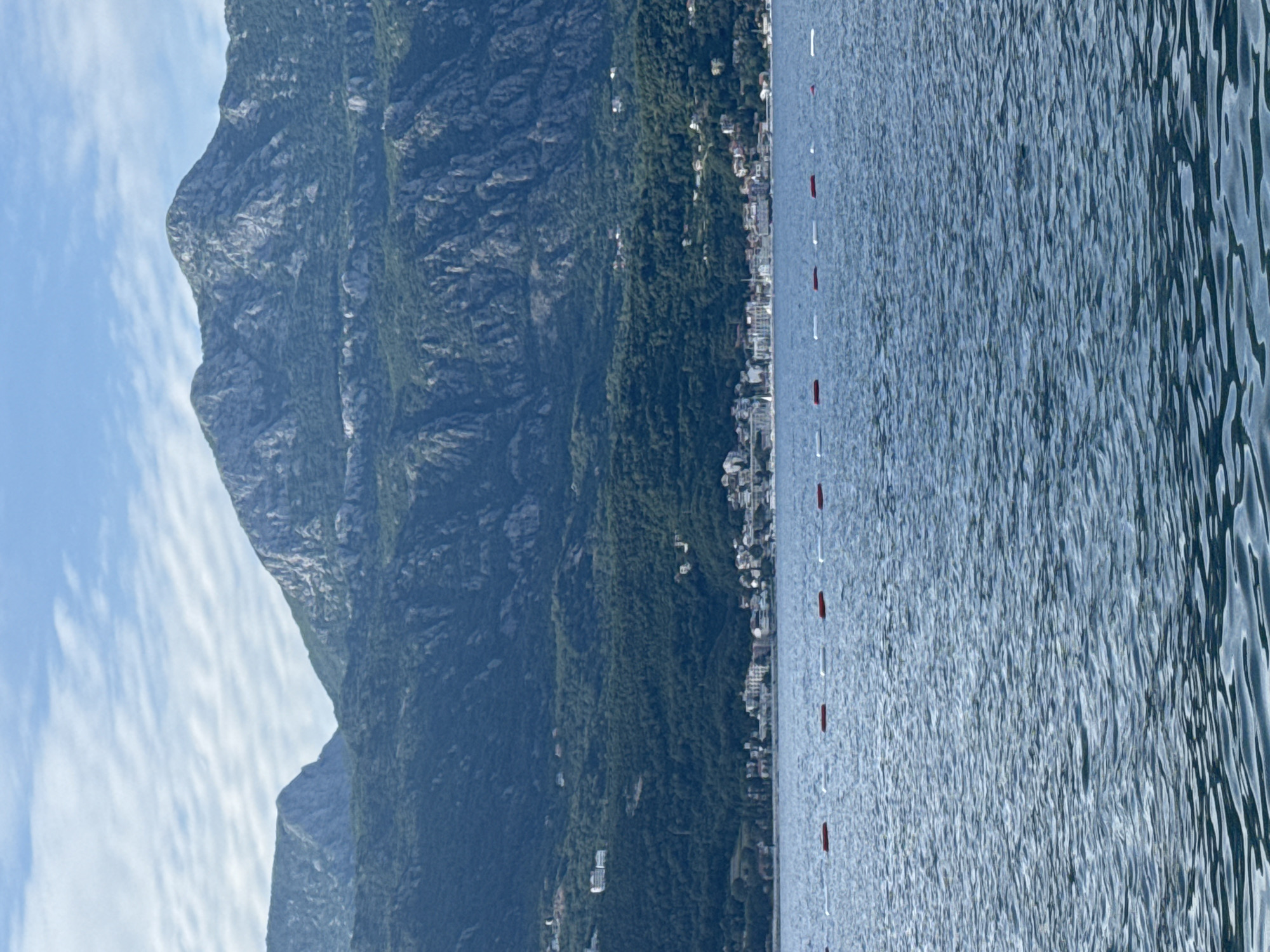 This is a vertical landscape photograph depicting a expansive body of water in the foreground, a coastal town at the base of large mountains in the mid-ground, and a partly cloudy sky overhead.
The top approximately one-quarter of the image shows a light blue sky with numerous thin, horizontally stretched white clouds, consistent with altocumulus or stratocumulus formations.
Below the sky, a series of rugged, multi-layered mountains dominates the mid-ground. These mountains are predominantly covered in dark green vegetation, with significant exposed grey rock formations and craggy peaks. Scattered buildings, primarily white, are visible amidst the greenery on the lower slopes of the mountains.
At the very base of the mountains, bordering the water, is a densely packed town or settlement composed of numerous light-colored buildings with some red-tiled roofs. The town stretches horizontally along the coastline. Details of boats or harbor infrastructure are faintly discernible within the town's water-frontage.
The bottom approximately two-thirds of the image is filled by a body of deep blue water, showing numerous small ripples and waves on its surface. Stretching horizontally across the mid-foreground of the water is a series of uniformly spaced, alternating red and white cylindrical buoys, delineating a boundary or zone. There are six visible red buoys and six visible white buoys.
No people are visible in the scene. The lighting suggests daytime conditions. The specific location within "Unknown, Unknown" is not identifiable from the visual content.