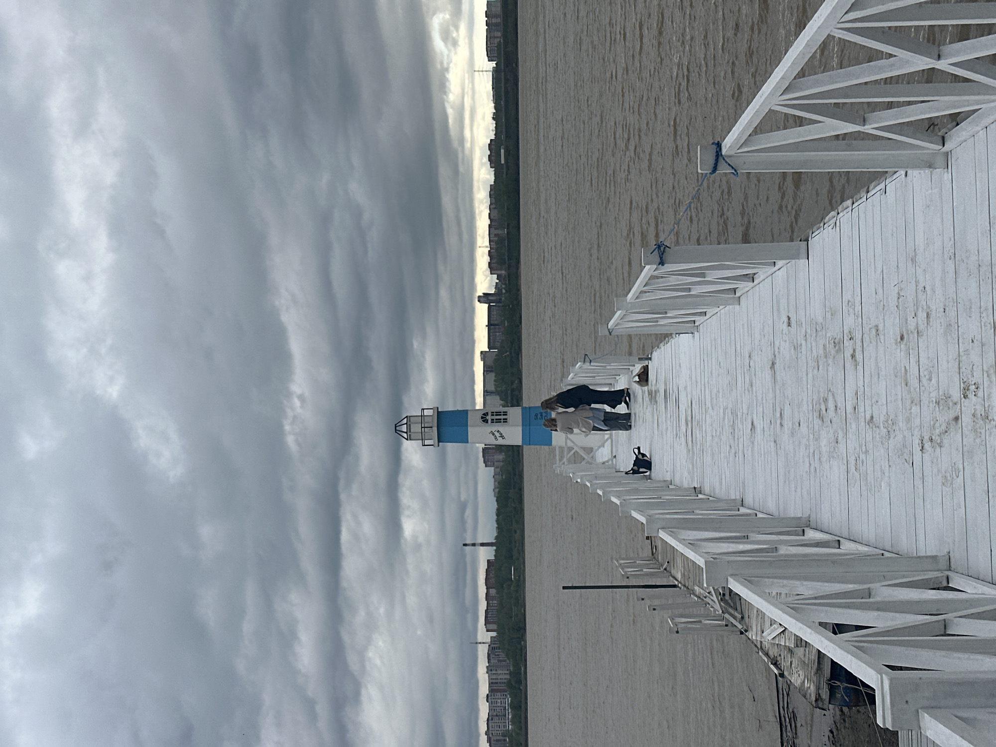 The image depicts a pier extending into a body of water, with a structure resembling a lighthouse on the pier. The sky is overcast with dense clouds. A shoreline with buildings, including tall apartment blocks and what appears to be a smokestack, is visible in the background, likely indicating an urban area. Two individuals appear to be standing near the lighthouse structure on the pier. The pier is constructed of wood and painted white. The scene suggests the pier is located in Berdsk, Russia.