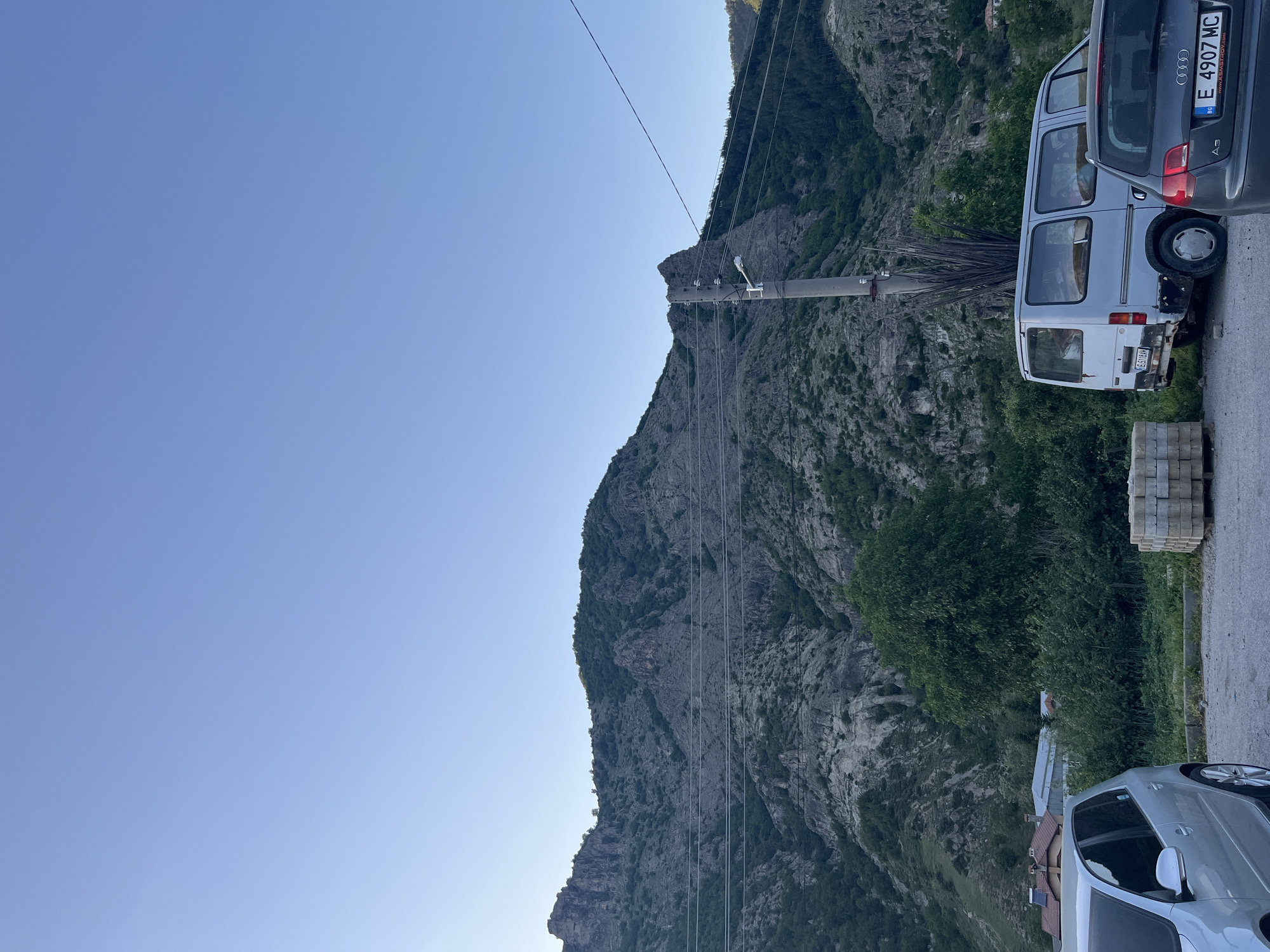 The image shows a mountainous landscape under a clear blue sky. A large rocky mountain dominates the center of the frame, with visible power lines running along its slope. In the foreground, on the right side of the image, are two parked vehicles: a white van and a grey hatchback. Adjacent to the vehicles, there is a stack of paving stones on the side of the road. The image is taken at Bukovo, Bulgaria.