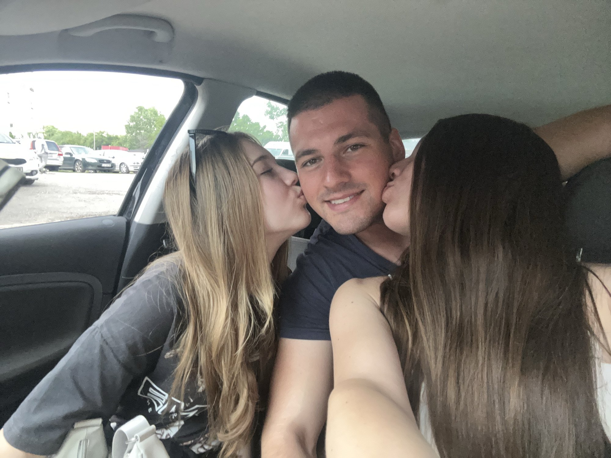 Inside a vehicle, one male and two females are depicted in a selfie. The male, positioned centrally in what appears to be the driver's seat, has short dark hair and is smiling directly at the camera. A female with long blonde hair, wearing a dark grey t-shirt with a white graphic and sunglasses perched on her head, is kissing the male's left cheek. Another female with long dark brown hair, wearing a white top, is simultaneously kissing the male's right cheek. The car interior shows a grey headliner and dark door panels. Through the car windows, an outdoor parking lot is visible under an overcast sky, containing several parked vehicles including a dark green sedan with a partially visible Bulgarian license plate displaying "CA 55", indicating the city of Sofia, Bulgaria. Green foliage from trees is present in the distant background.