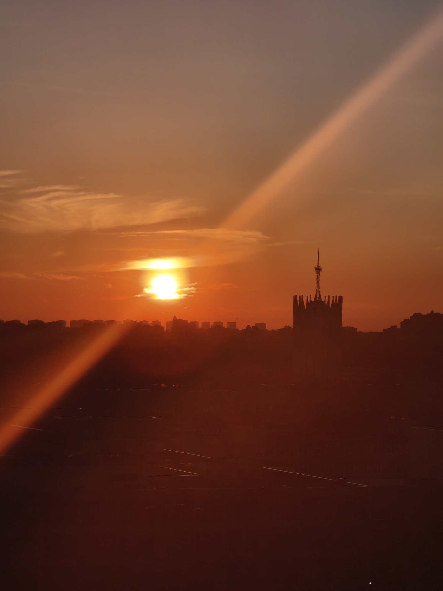 The image depicts a sunset over Saint Petersburg, Russia. The sun is a bright white orb in the center of the frame, with orange light radiating outwards. A prominent light beam stretches diagonally across the image. The cityscape is silhouetted against the bright sky. In the middle ground is a large, ornate building with a tall spire, likely a landmark structure in Saint Petersburg. The bottom of the image is darker, obscuring details of the foreground.