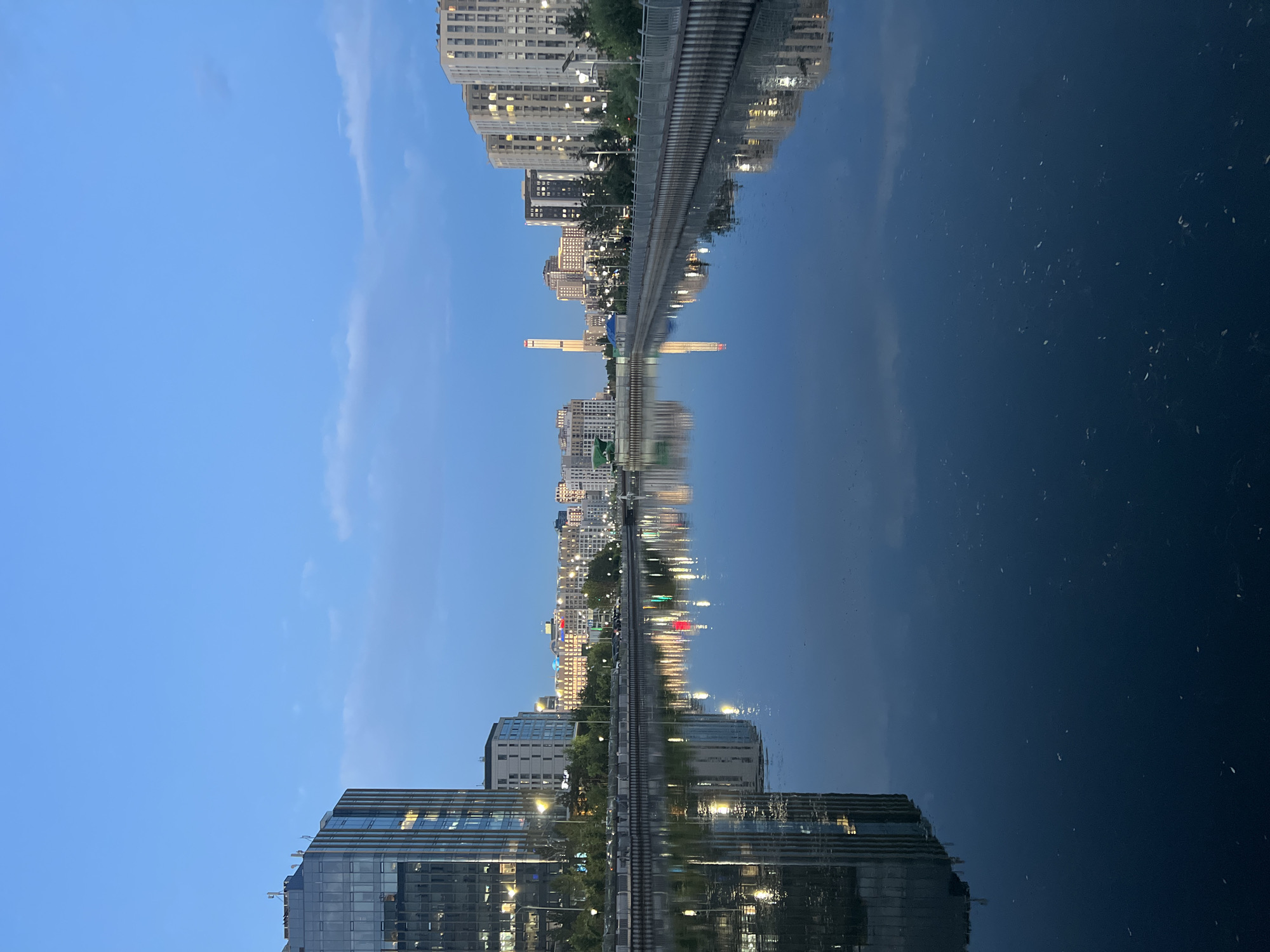 This is a horizontal, eye-level, medium shot of the Ishim River in Astana, Kazakhstan, at dusk. On the left, a tall, modern glass-fronted office building reflects in the water. Across the river, a row of residential buildings lines the embankment, with lights visible in the windows. Beyond them, a collection of taller, modern buildings form the city skyline, including a distinctive, slender tower illuminated at the top. The water is calm and dark, reflecting the buildings and the sky, which is a deep blue with scattered clouds. A metal fence runs along the embankment, separating the water from the buildings and street. The scene is devoid of people.