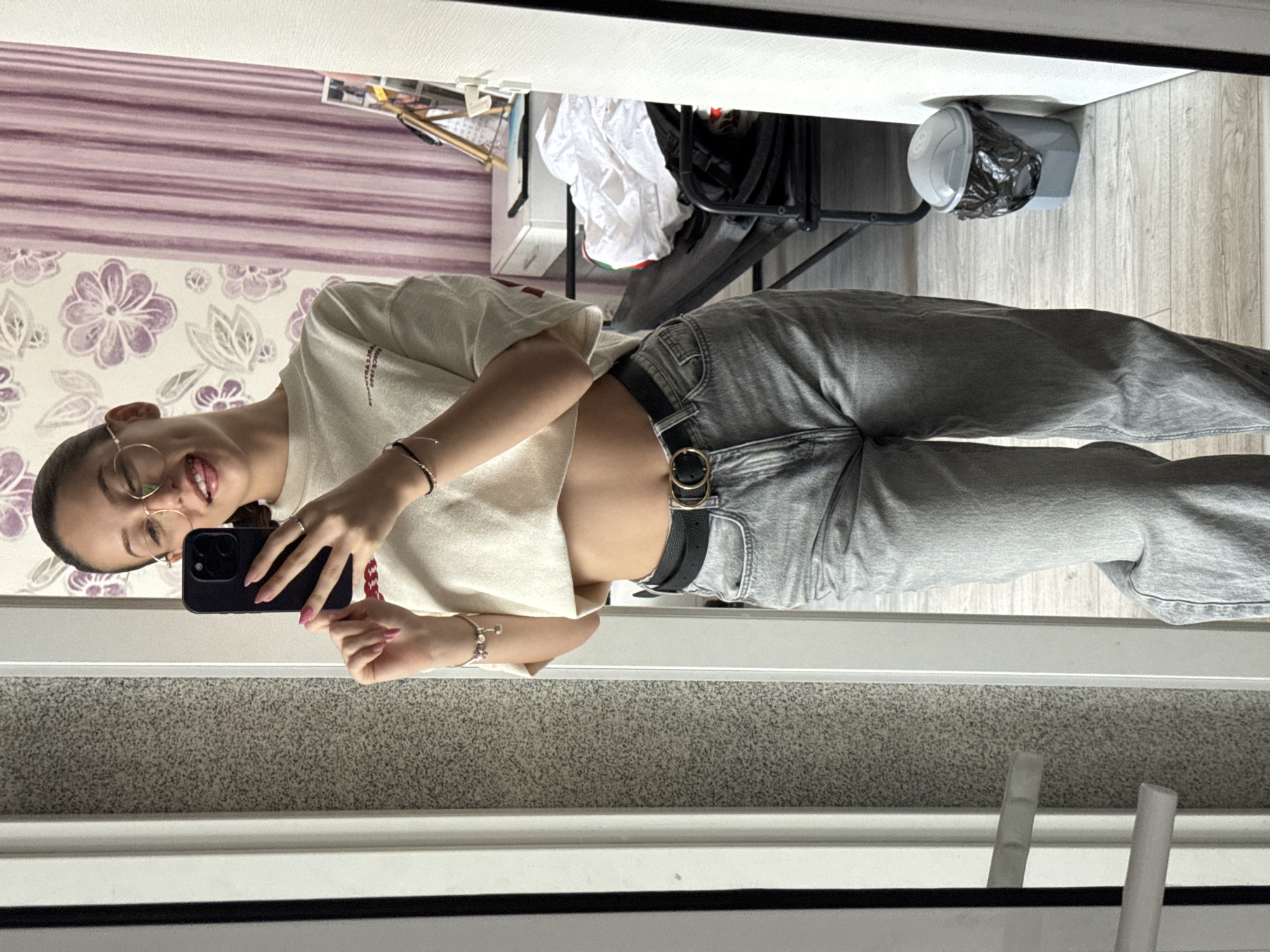 A young woman with dark hair pulled back and wearing glasses is taking a selfie in a mirror. She has on a light-colored, cropped t-shirt and light-wash denim jeans with a black belt. Her tongue is sticking out slightly, and she is holding a black smartphone in her right hand. Her left hand is visible, showing pink fingernails and a bracelet. The background includes a wall with floral wallpaper and vertical purple stripes, a desk with items on it, a chair, and a trash can. The floor is made of light-colored wood planks. This scene appears to be indoors, possibly in a residential setting in Sofia, Bulgaria, though no specific Sofia landmarks are identifiable.