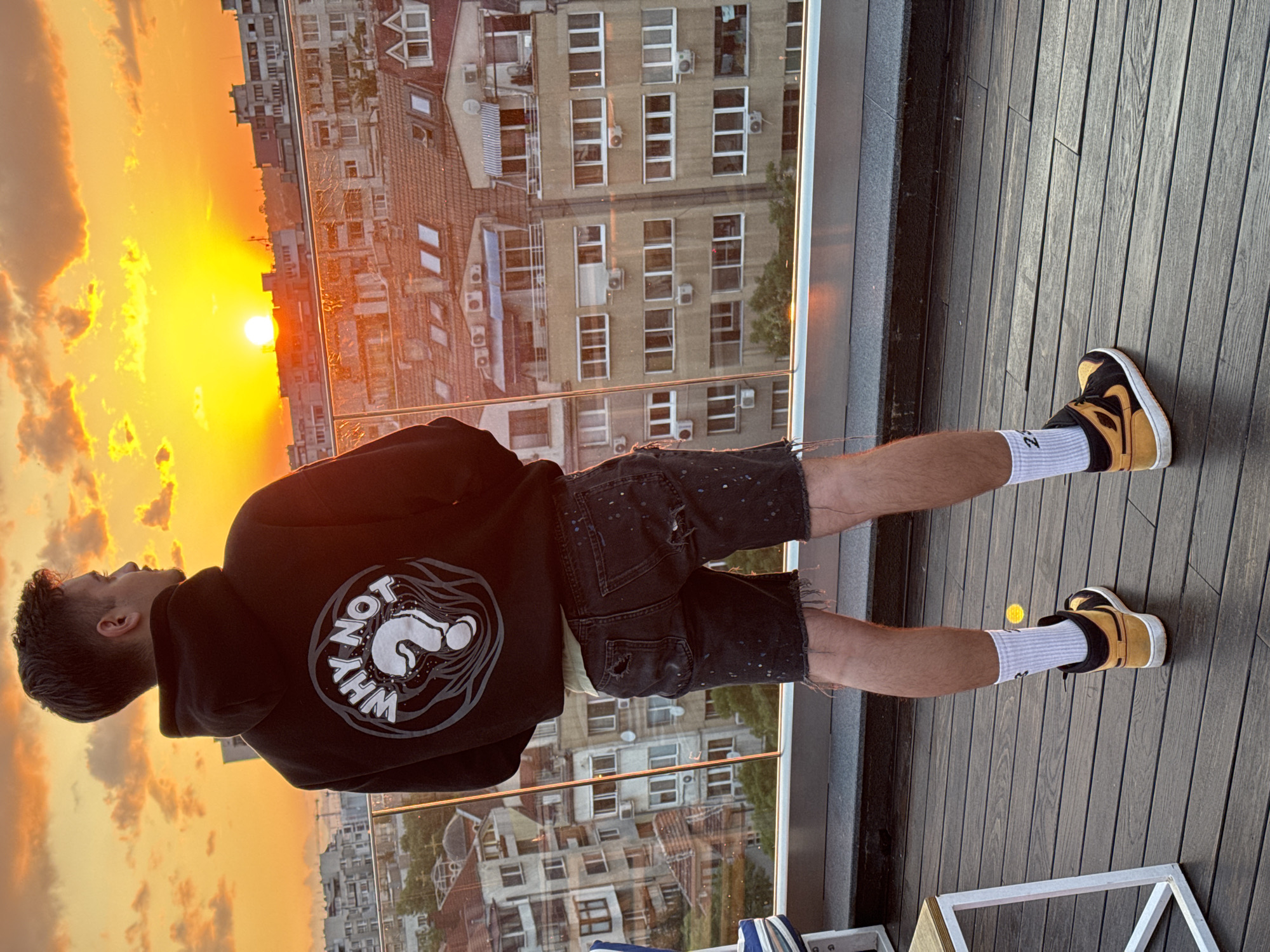 A male individual stands on a wooden deck, viewed from behind, looking towards a sunset over a cityscape. He wears a black hooded sweatshirt with a white circular graphic on the back that reads "WHY NOT" surrounding a prominent question mark. Beneath the hoodie, he wears distressed black denim shorts with white paint splatters and frayed hems. He has white crew socks, one of which displays the number "23" in black, paired with black and yellow high-top sneakers. A glass railing separates the deck from the extensive urban environment. The background features numerous multi-story buildings, typical of residential or mixed-use blocks, under a vivid orange and yellow sky. The sun is visible as a bright, low-lying disc near the horizon, bathing the scene in warm light. The architecture of the buildings, including some with red tile roofs and visible air conditioning units, is characteristic of the city of Sofia, Bulgaria.