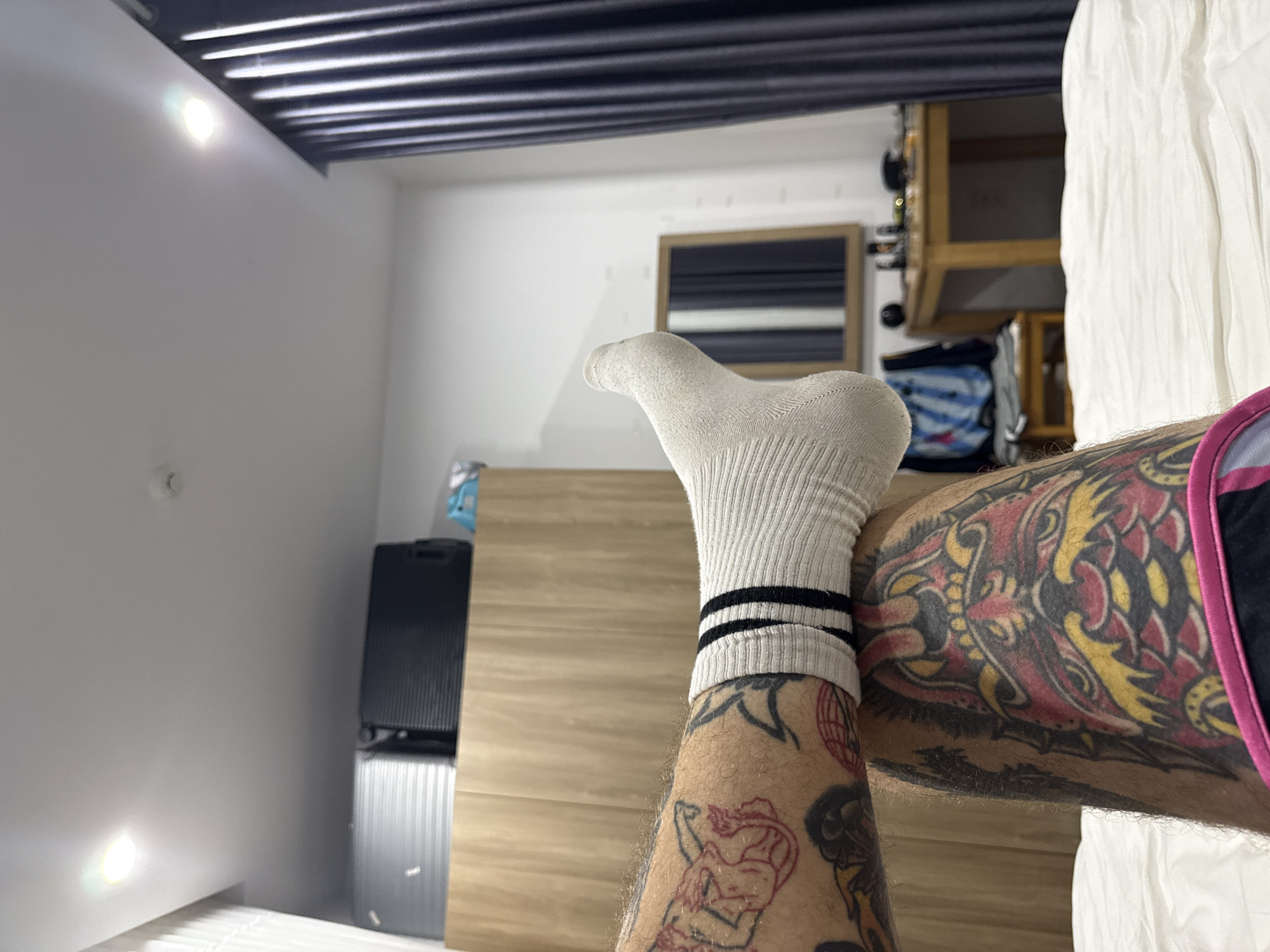 The image shows a person's leg and foot extended toward the camera. The foot is covered with a white sock with two black stripes near the ankle. The leg has multiple tattoos. A portion of the leg shows a design in red ink of a figure with a cowboy hat. The thigh has a large, colorful tattoo of a dragon with red, yellow, and black ink. The person is wearing black athletic shorts with a pink stripe along the edge. The background shows a room with a white wall on the left, a black suitcase or cabinet, a light wood cabinet, a mirror, and shelves of miscellaneous items on the upper right. The ceiling is dark with parallel lines, potentially a slat ceiling or overhead lighting fixture. The location is not identifiable as Đà Nẵng, Vietnam based on the available visual information.