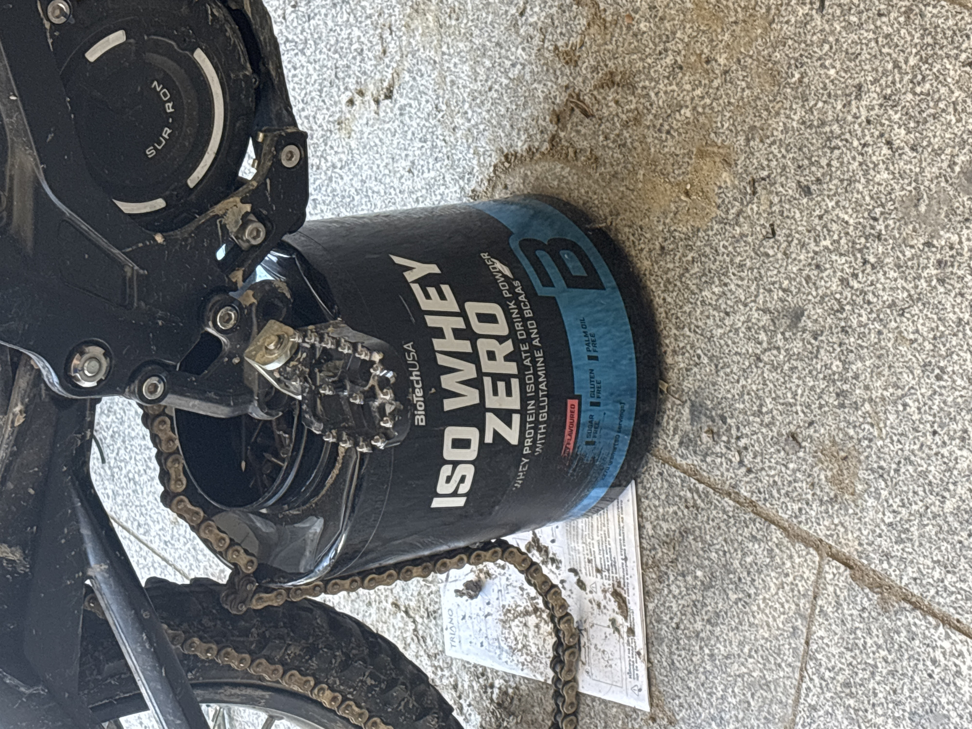 The image shows a Sur-Ron electric bike leaning against a large black and blue container of BiotechUSA ISO Whey Zero protein powder. The bike, notably dirty, has a visible chain, pedals, and the Sur-Ron logo on the motor cover. A partially visible document is under the protein container, with the word "TRIONO" visible on it. The scene is set on a grey tiled surface with some dirt and debris. There is no indication of the location of the city of Asenovgrad, Bulgaria.