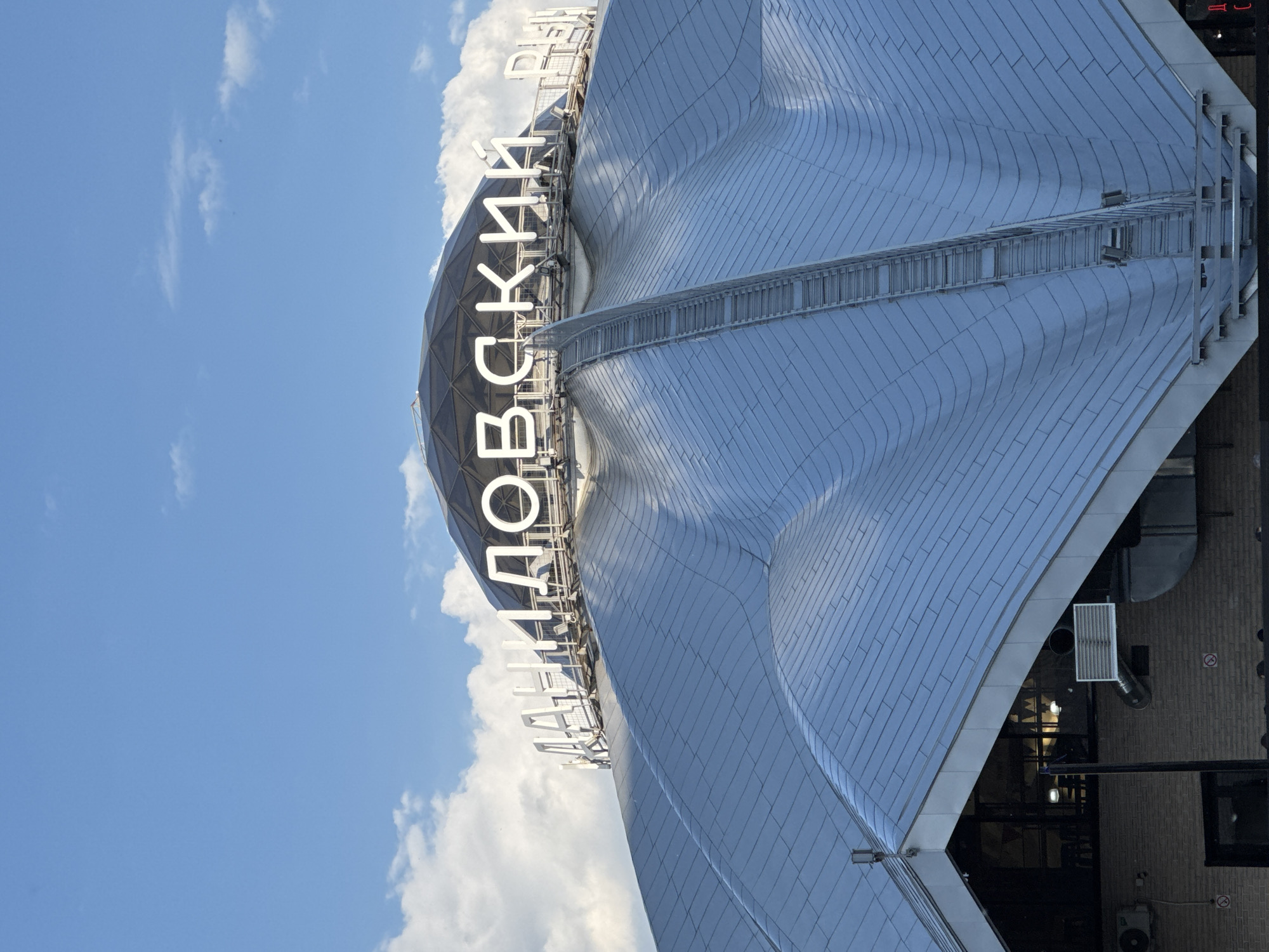 The image displays a large, modern architectural structure, likely a stadium or sports complex, set against a bright blue sky with scattered white clouds. The building's most prominent feature is its distinctive, undulating roof, composed of numerous metallic, possibly silver or light gray, panels that create a wavy texture. A long, ladder-like maintenance walkway is visible traversing the curves of the roof.
Prominently displayed on the upper part of the structure are large, white, illuminated Cyrillic letters. The clearly visible text spells out "ЛУЖНИКИЙ" (Luzhnikiy), indicating a connection to the Luzhniki Stadium, a well-known sports arena in Moscow, Russia, which aligns with the provided location context.
The scene appears to be captured during daytime under clear weather conditions, as suggested by the bright sky and crisp lighting on the building's surface. In the lower right corner of the frame, glimpses of adjacent building elements are visible, including dark glass or openings and a textured wall, possibly made of brick, along with some industrial-looking vents or equipment. No people or activities are depicted; the focus is solely on the building's striking architecture. The overall impression is one of modern design and scale.