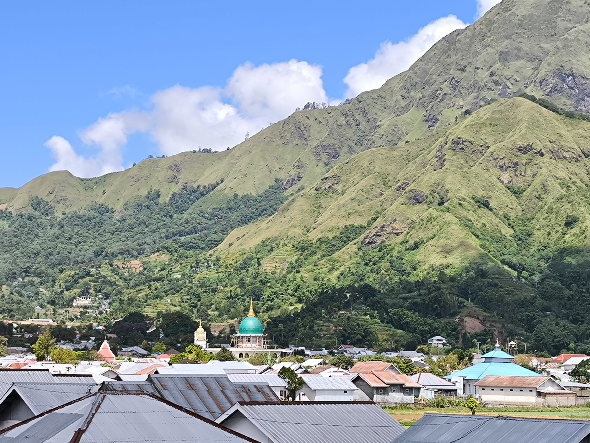 The image captures a wide-angle view of Sembalun Bumbung, Indonesia, dominated by a backdrop of verdant, rolling mountains under a bright blue sky with scattered white clouds. In the foreground, the rooftops of densely packed houses with primarily grey and some red tiling are visible, presenting an aerial perspective.
Mid-ground features include a mosque distinguished by its green dome and a smaller structure with a blue dome. These buildings indicate religious and community spaces within the town. Patches of green fields and trees are interspersed between the houses, suggesting an agrarian landscape.
The scene appears to be shot during the daytime, with good visibility. No people are directly visible within the frame. The overall impression is of a rural Indonesian town nestled in a mountainous region.