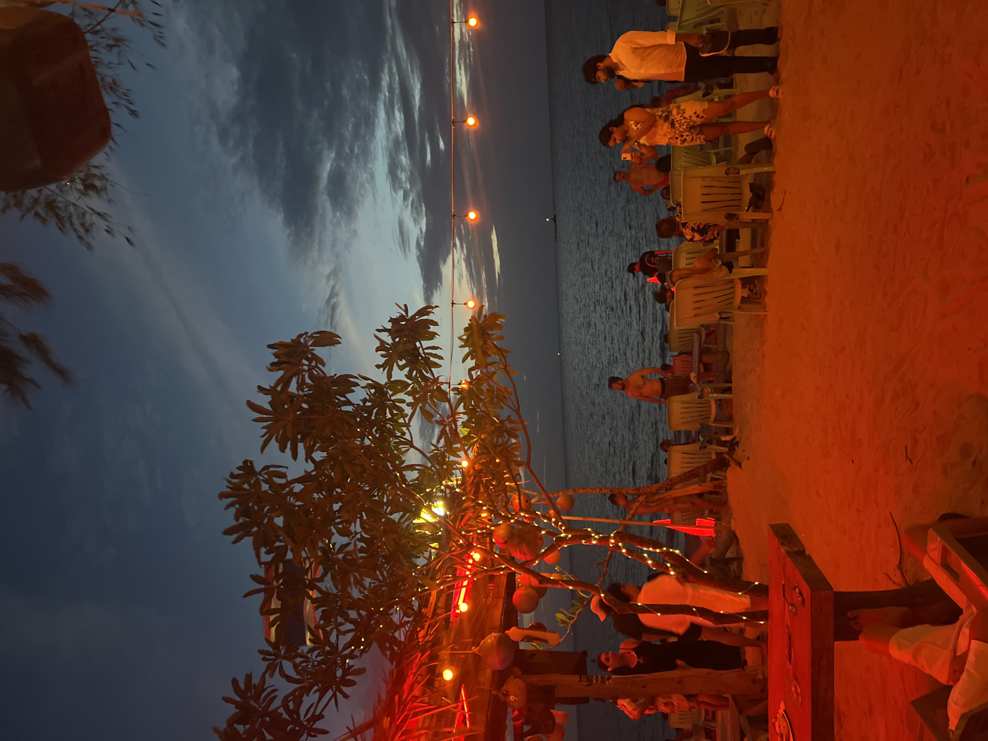 At dusk on a sandy beach in Ko Pha-ngan District, Thailand, a group of people are gathered at an open-air establishment. A thatched-roof structure with decorative lights and hanging objects is visible on the left, illuminated by red lighting. String lights are draped on a tree in front of the structure, and more lights are strung across the scene above the beachgoers.
Several individuals are seated on plastic chairs arranged on the sand, facing the ocean. Some are standing and interacting, with one person taking a photo with a smartphone and another holding a microphone. A man is standing shirtless near the water's edge, looking out at the sea. The ocean is visible in the background, with a calm surface and a distant light on the horizon. The sky is a deep blue, transitioning from twilight to night, with scattered clouds. The red lighting from the establishment casts a warm glow on the sand and the people, creating a festive atmosphere.