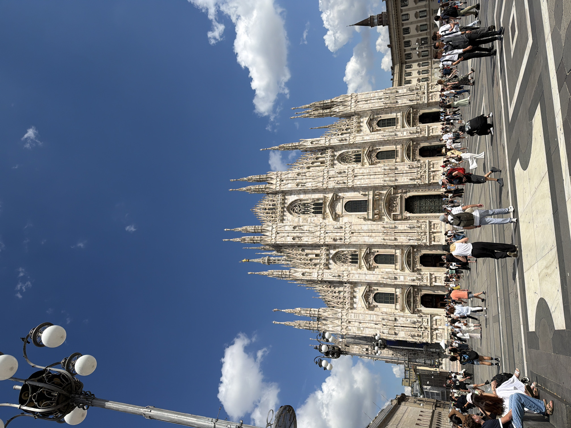 An outdoor, wide-angle shot captures the grandeur of the Milan Duomo and the bustling Piazza del Duomo under a clear blue sky. The magnificent Gothic cathedral, located in Milan, Italy, dominates the background, showcasing its intricate white marble facade, countless spires, ornate statues, and decorative windows. Its impressive scale is emphasized by the detailed architectural elements visible across its multiple levels.
In the mid-ground and foreground, a large, expansive public square is filled with numerous people. The square features a distinctive paving pattern of alternating dark and light stripes or blocks, guiding the eye through the scene. People of various ages and ethnicities are seen walking, standing, and observing the cathedral. They are dressed in casual attire, consistent with warm weather, including t-shirts, shorts, dresses, and jeans. Some individuals appear to be tourists sightseeing, while others are simply moving through the square. In the lower right corner, a few people are seated on the ground, relaxing. A group of men in suits or vests is also visible, perhaps part of a tour group or performing.
To the far right, other historical city buildings with traditional architecture are visible, and a distant spire rises against the sky. In the lower left foreground, a decorative street lamp with multiple spherical white lights adds an urban detail to the composition.
The time of day appears to be sunny daytime, with a bright blue sky dotted with scattered white cumulus clouds, indicating pleasant weather. The overall atmosphere is one of a vibrant, active public space at a renowned historical landmark. No visible text is discernible in the image.