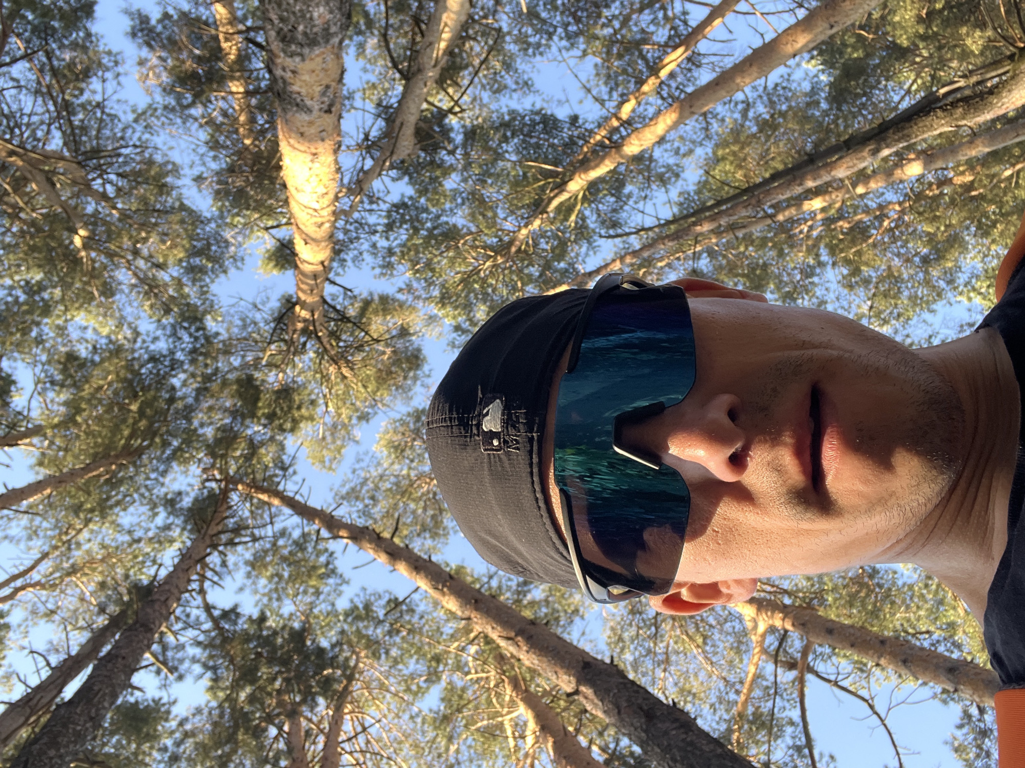 A low-angle photograph features a man wearing sunglasses and a black cap, with a background of trees and blue sky. The man's face is partially illuminated by sunlight. The sunglasses have a reflective surface, and the trees appear to be pine trees. The location is most likely in a forest in Chaikovsky, Russia.