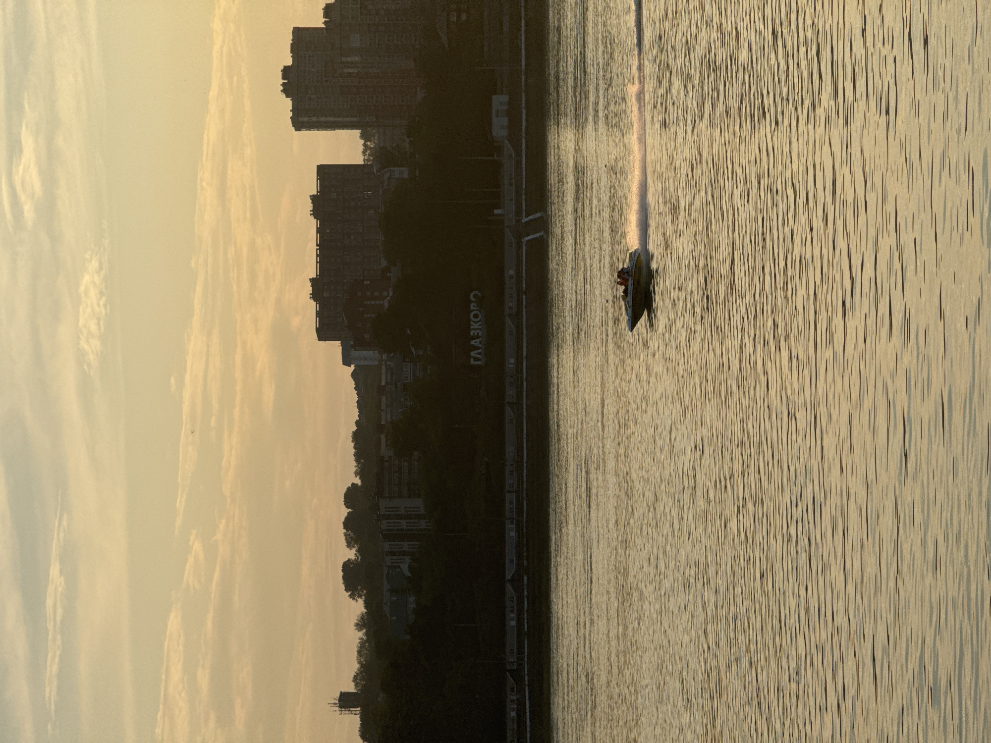 The image depicts a scenic view of the Angara River in Irkutsk, Russia, at sunset. The sky is a soft, golden hue, with streaks of pale clouds. On the far bank, a tree-lined embankment separates the river from the city skyline. Silhouetted against the sky are several tall, modern apartment buildings. Below these buildings, a sign with Cyrillic lettering "ГЛАЗКОРО" is visible. In the foreground, a motorboat with two occupants speeds across the water, leaving a white wake behind it. The river's surface is rippled, reflecting the warm light of the setting sun.