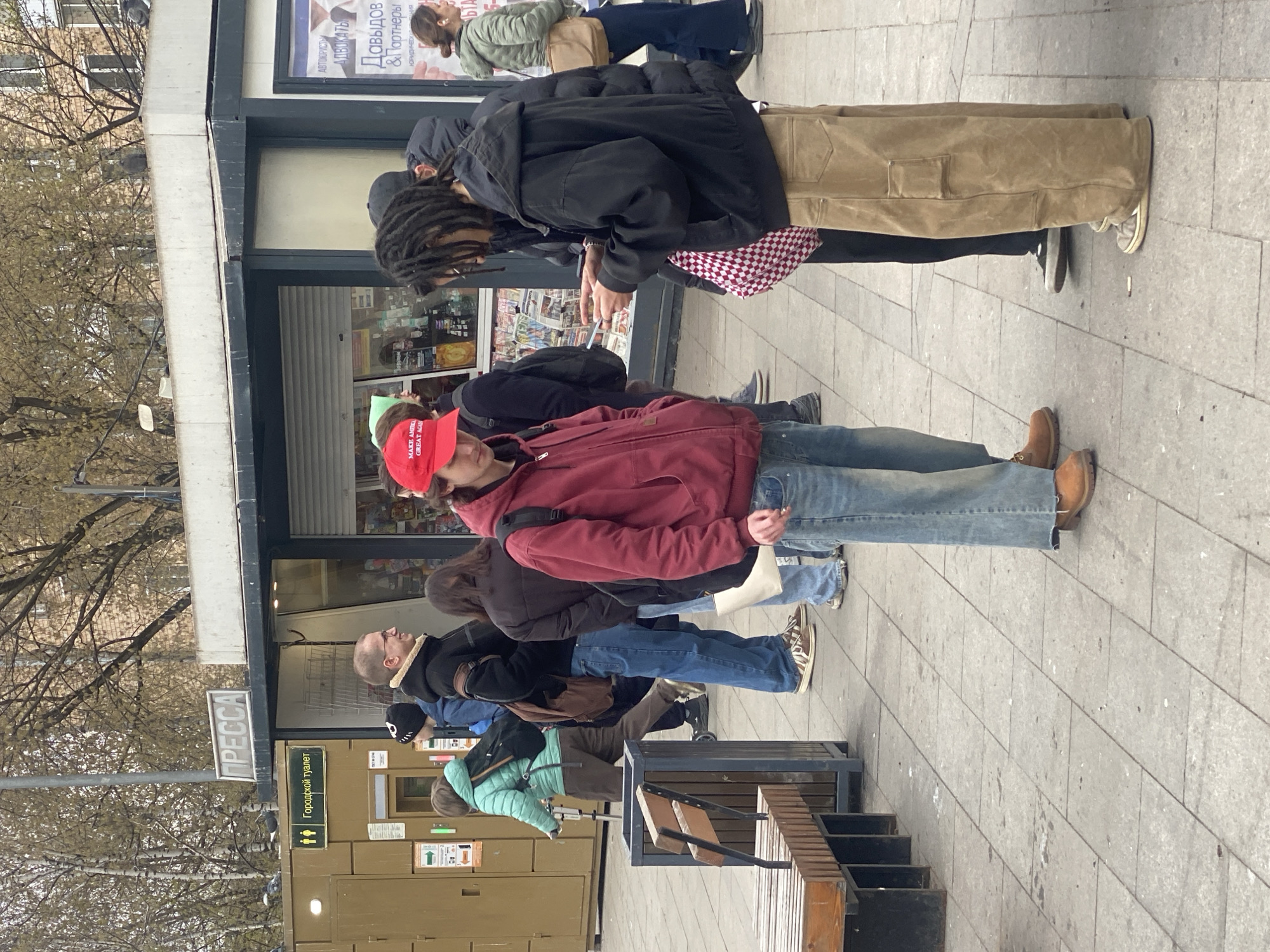 The image captures a scene in Moscow, Russia, where people are standing in a line in front of a kiosk. The kiosk has a sign that reads "ПРЕССА" (Press) and displays the words "Городской туалет" (City Toilet). Several people are visible, including an individual wearing a red "MAKE AMERICA GREAT AGAIN" hat. The kiosk appears to sell newspapers or magazines. Additionally, there's a sign referring to legal services ("АВТОЮРИСТЫ АДВОКАТЫ" and "Давыдов & Партнеры"). The image also shows the edge of a bench and the pavement. The trees in the background show that it is likely springtime.