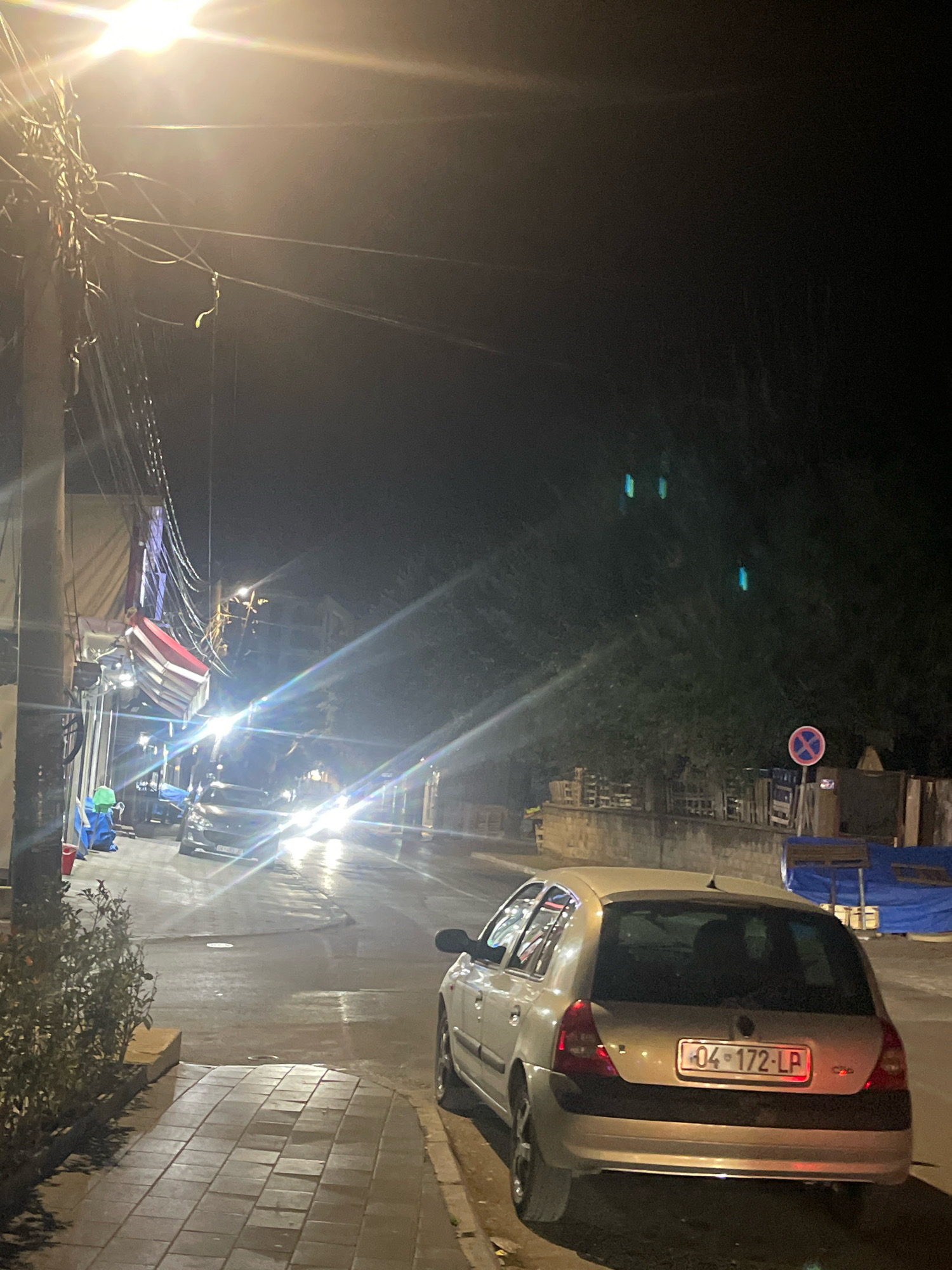The image captures a nighttime street scene in Prizren, Kosovo, characterized by artificial illumination and wet pavement. A silver hatchback car, license plate "04-172-LP", is parked on the right side of the road, facing away from the viewer. Its taillights are illuminated. The street appears to be paved with asphalt, and the sidewalk to the left is made of rectangular stone tiles.
On the left side of the street, a collection of items is visible, including blue tarps, a green bin, and a red bucket. Overhead, a dense network of electrical wires and poles spans the street, with a bright streetlamp casting a strong glare. In the background, buildings are dimly visible, and trees line the right side of the road. A circular traffic sign with a red circle and a diagonal red bar is posted on the right. The overall impression is of a quiet urban environment at night, possibly after rainfall.