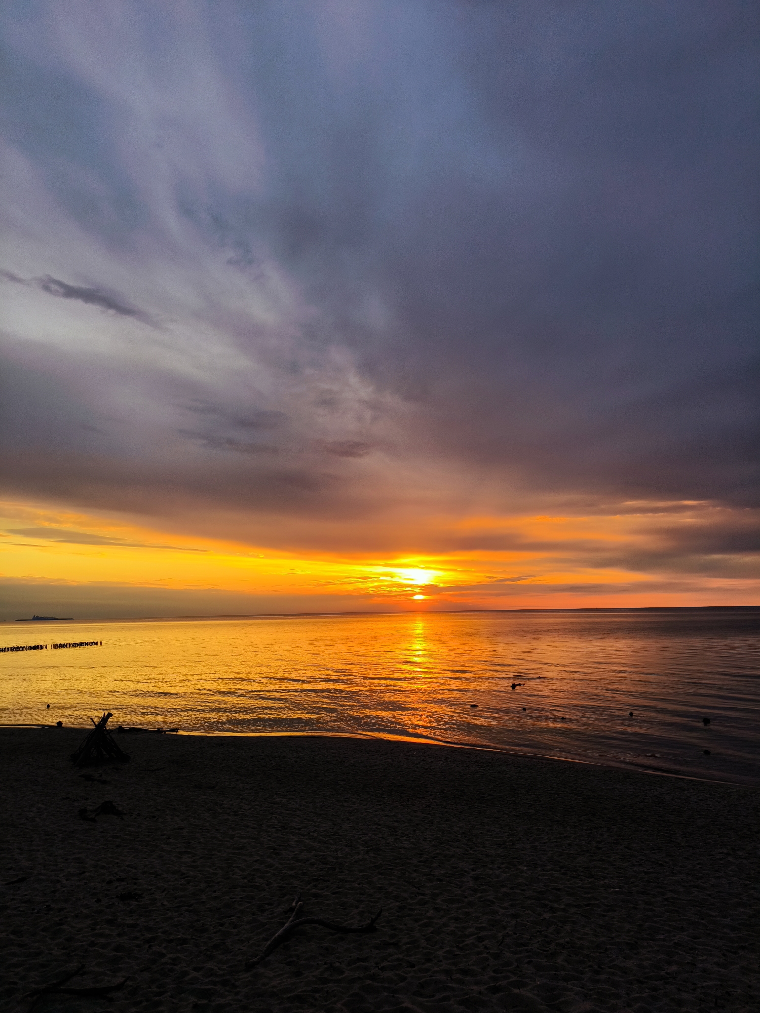 The image depicts a sunset over the Baltic Sea from a sandy beach in Zelenogradsk, Russia. The scene is devoid of human presence.
The upper approximately two-fifths of the frame is dominated by a dramatic sky. The uppermost section features heavy, dark purplish-grey and deep blue clouds, with some lighter grey streaks indicating movement and varying thickness. Closer to the horizon, the sky transitions into a vibrant band of intense golden-orange and yellow, where the sun is visible as a distinct, bright orb slightly to the left of the central vertical axis, situated directly on the horizon line. Clouds in this lower sky section appear as layered strata in shades of orange and muted purplish-grey, partially diffusing the intense light.
The middle section of the image, approximately two-fifths, is occupied by the sea. The water directly beneath the sun and extending towards the foreground reflects the sun's glow intensely, appearing brilliant golden-orange with visible ripples. Towards the right and further from the direct reflection, the water color deepens to a reddish-brown. On the far left, a line of dark, vertical wooden poles, consistent with groynes or breakwaters, extends into the water from an unseen shore. A faint, dark silhouette of a distant landmass or large structure is discernible on the far left horizon. Several small, dark, irregular objects, possibly rocks or debris, are scattered on the water surface in the mid-ground.
The bottom approximately one-fifth of the image consists of a dark, sandy beach in the foreground. The sand is heavily shadowed, appearing almost black or deep brown, contrasting sharply with the brightly lit water and sky. The texture of the sand is subtly visible. On the left side of the beach, a small, conical structure constructed from dark wood branches, resembling a teepee, is present. A few individual pieces of dark driftwood are scattered on the sand in various locations across the foreground.