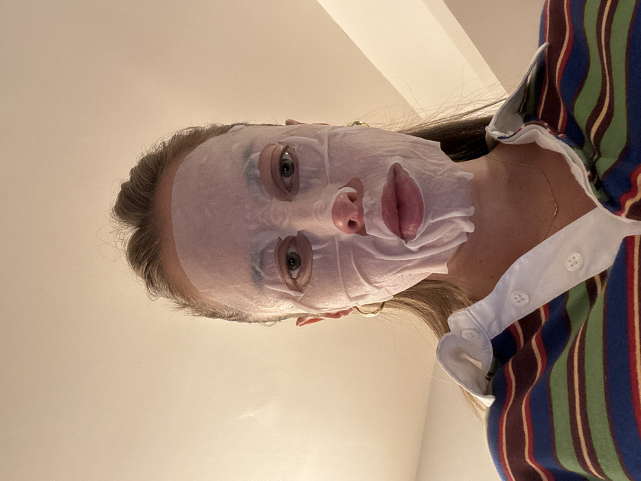 A person wears a white sheet mask on their face. The mask covers the forehead, nose, cheeks, and lips, leaving openings for the eyes. The person has fair skin, light brown hair, and is wearing a striped rugby shirt with a white collar. They are facing upward, looking at the camera. The background is a plain off-white wall. Gold hoop earrings are visible.