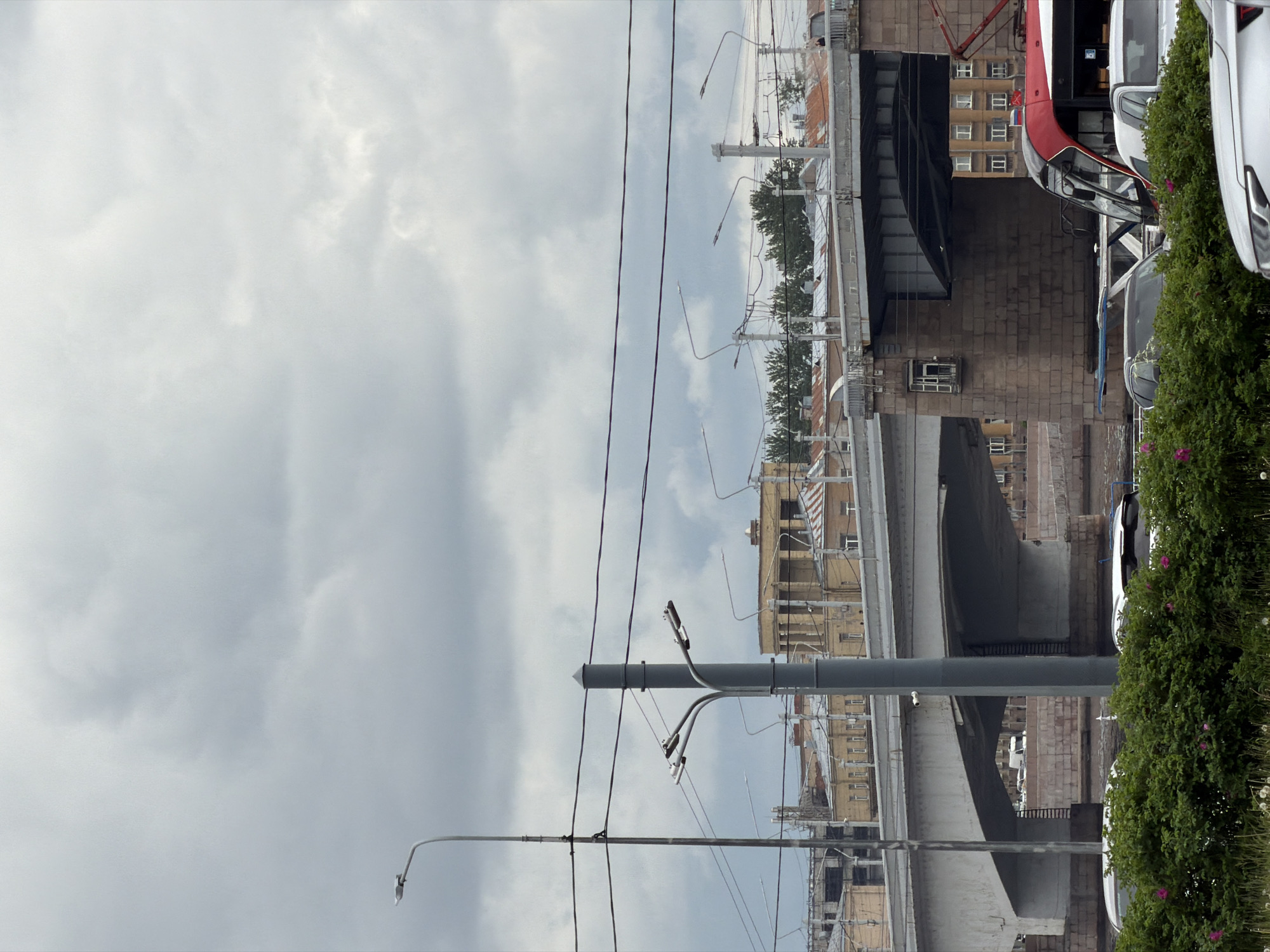 The image shows a view of the Novo-Kamenny Bridge in Saint Petersburg, Russia, from below. The underside of the bridge is visible, along with utility poles, power lines, and streetlights. Buildings are visible in the background. Parked cars and greenery are seen on the side of the bridge. The sky is overcast.