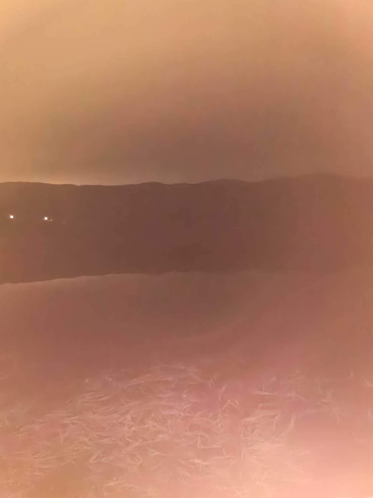 The image depicts a dusky, hazy landscape. In the background, a dark band of hills or mountains is visible against a lighter sky. A lower, snow-covered plain extends towards the foreground. The ground appears textured with lines and patterns, suggesting ice or frozen vegetation. Several small lights are present to the left of the image. The location is Unknown, Unknown.