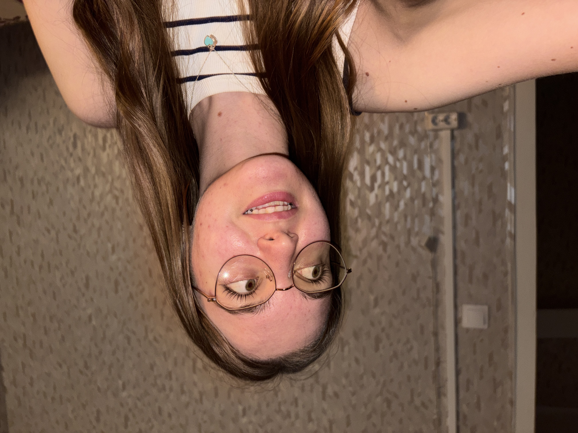 A young woman with long brown hair is shown upside down in a medium shot. She wears round, wire-rimmed glasses and a white and blue striped tank top with a necklace. The woman looks up and to the side with a slight smile. The background features a beige textured wall with a light switch and part of a door frame visible. It is impossible to determine if the image was taken in the city of Perm, Russia, or elsewhere.