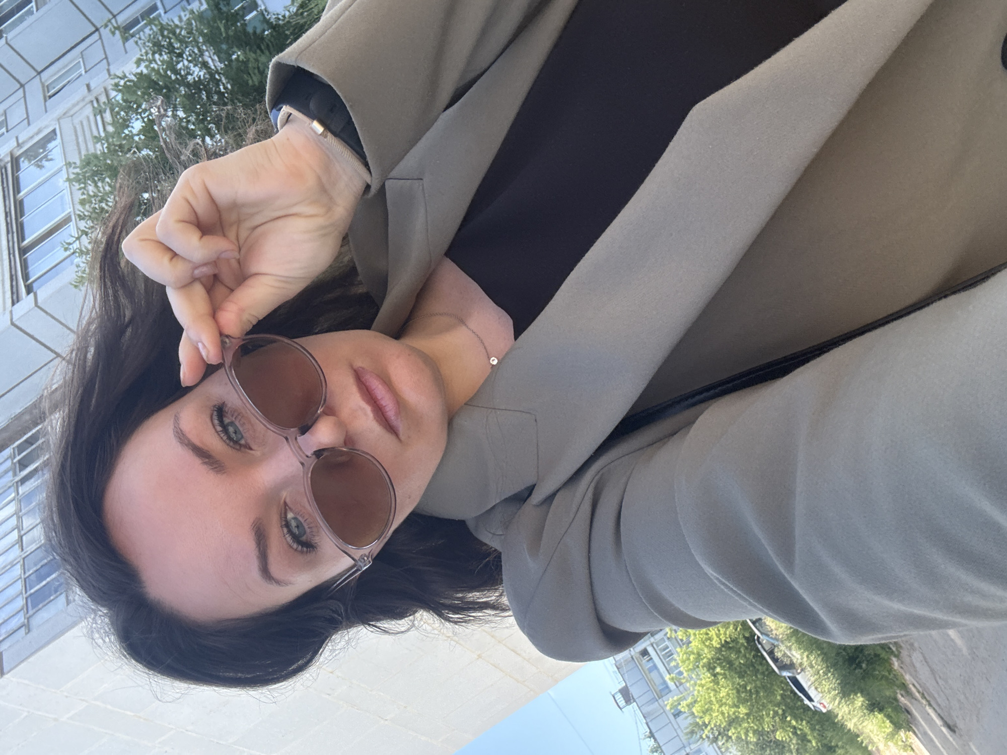 A medium shot captured from a slightly low angle shows a woman with dark hair and fair skin posing for a selfie outdoors. She is wearing oversized sunglasses with brown lenses, a light grey blazer over a dark top, and a thin silver necklace with a small pendant. Her right hand is raised, adjusting the sunglasses. The background features a modern building with multiple windows and some greenery, suggesting an urban environment in Chaikovsky, Russia. The lighting indicates it is daytime.