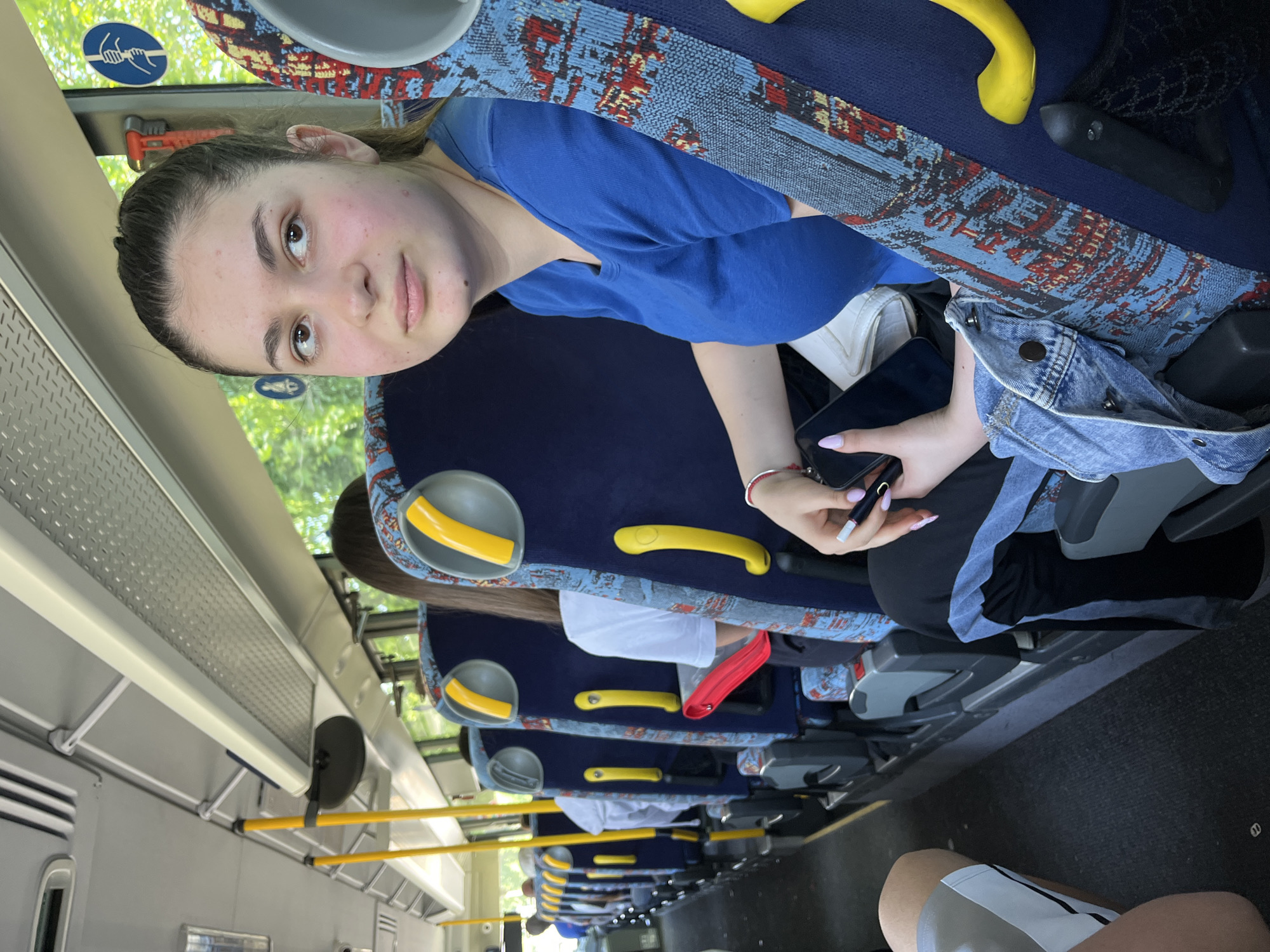 The image captures the interior of a public transport bus during the daytime. The main subject is a young woman, likely in her late teens or early twenties, seated upright on the left side of the bus. She has light skin, dark hair pulled back, and dark eyes, looking directly at the camera with a neutral expression. She wears a blue t-shirt and a red bracelet on her right wrist. In her hands, she holds a black mobile phone and a small, black electronic device, possibly a vape pen. A white bag and a denim jacket are visible on the seat next to her.
The bus features blue seats with a distinct red and blue patterned fabric. Yellow handrails are prominently visible, attached to the backs of seats and along the overhead panels. Through the window adjacent to the main subject, green foliage is visible, indicating an outdoor, possibly natural, environment. An emergency hammer with a red handle is mounted on the window frame. A blue circular sticker, depicting a hand symbol and wavy lines, is also on the window, though specific text for instructions is illegible. Other passengers are partially visible further down the bus aisle. Overhead compartments and lighting fixtures line the bus ceiling, and the floor is dark. The scene suggests a casual journey or commute.