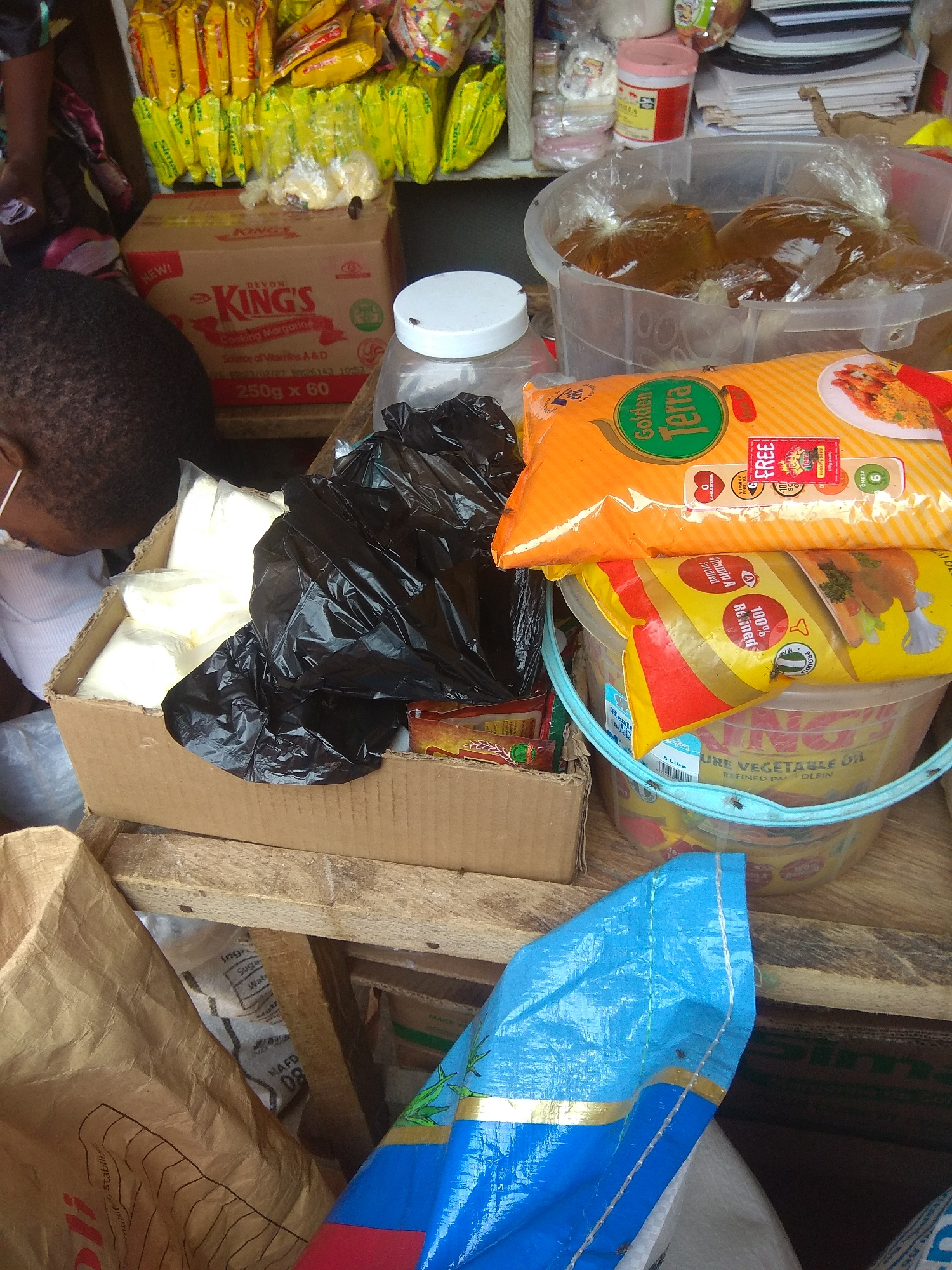 The image depicts a market stall in Jalingo, Nigeria, likely a roadside or open-air setup, with various food items and household goods. In the foreground, a person's head and shoulder are visible, with dark, textured hair and part of a white shirt. The person is looking downwards, suggesting engagement with the items on display.
Dominating the scene are several packaged goods. To the left, a brown cardboard box labeled "King's Cooking Margarine" is prominent, containing smaller packets. To the right of this box, a black plastic bag is filled with white packets, possibly food items like flour or sugar, and another black plastic bag appears to contain similar items.
Above these, a large, clear plastic tub holds multiple plastic bags filled with a dark brown liquid, likely cooking oil or a similar commodity. Resting on top of this tub are two brightly colored food packages. The top package is orange and green, labeled "Golden Terra", and features an image of shrimp. Beneath it is a yellow package with red stripes, also featuring food imagery.
Further back, shelves are laden with more yellow and red packaged goods, appearing to be snack items or condiments, typical of a Nigerian market. Several small plastic containers, one pink with a white lid, are also visible. The overall environment suggests a bustling commercial space with limited organization. The presence of flies on some of the items indicates potential hygiene concerns. The wooden structure supporting some of the items suggests a rustic and informal market setting.