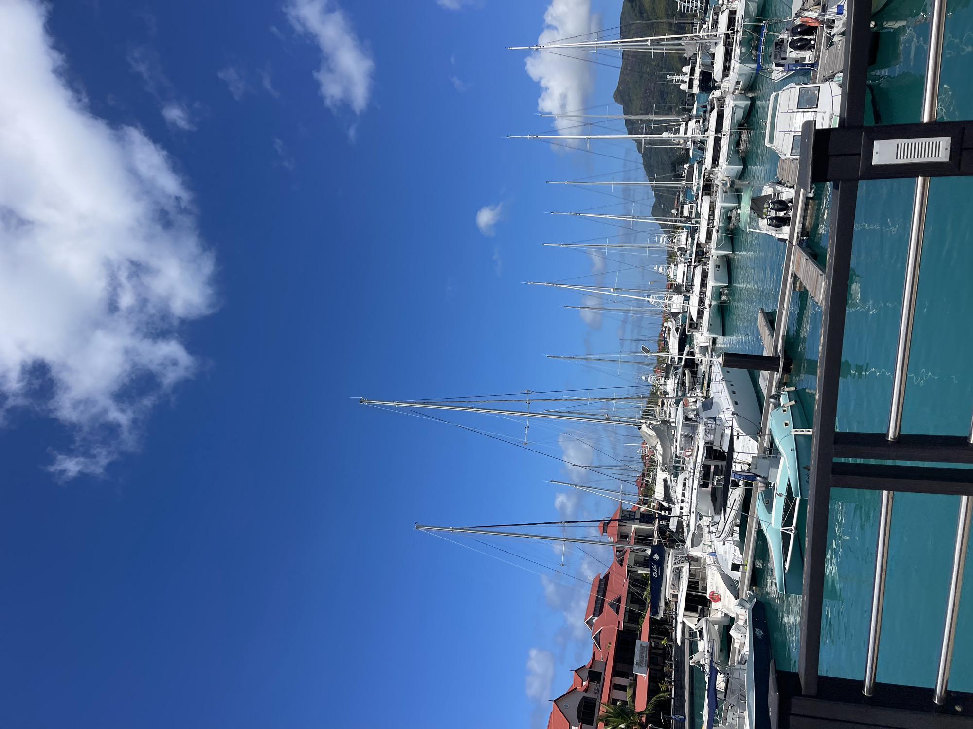 The image depicts a marina filled with numerous sailboats and motorboats under a bright blue sky with scattered clouds. In the background, several buildings with red roofs are visible, possibly indicating a waterfront development or hotel. The water in the marina is a clear turquoise color, reflecting the surrounding boats and the sky. The scene appears to be in a tropical or subtropical location, given the lush greenery on the distant hills and the style of architecture. It is likely a popular tourist destination or a well-established yachting hub.