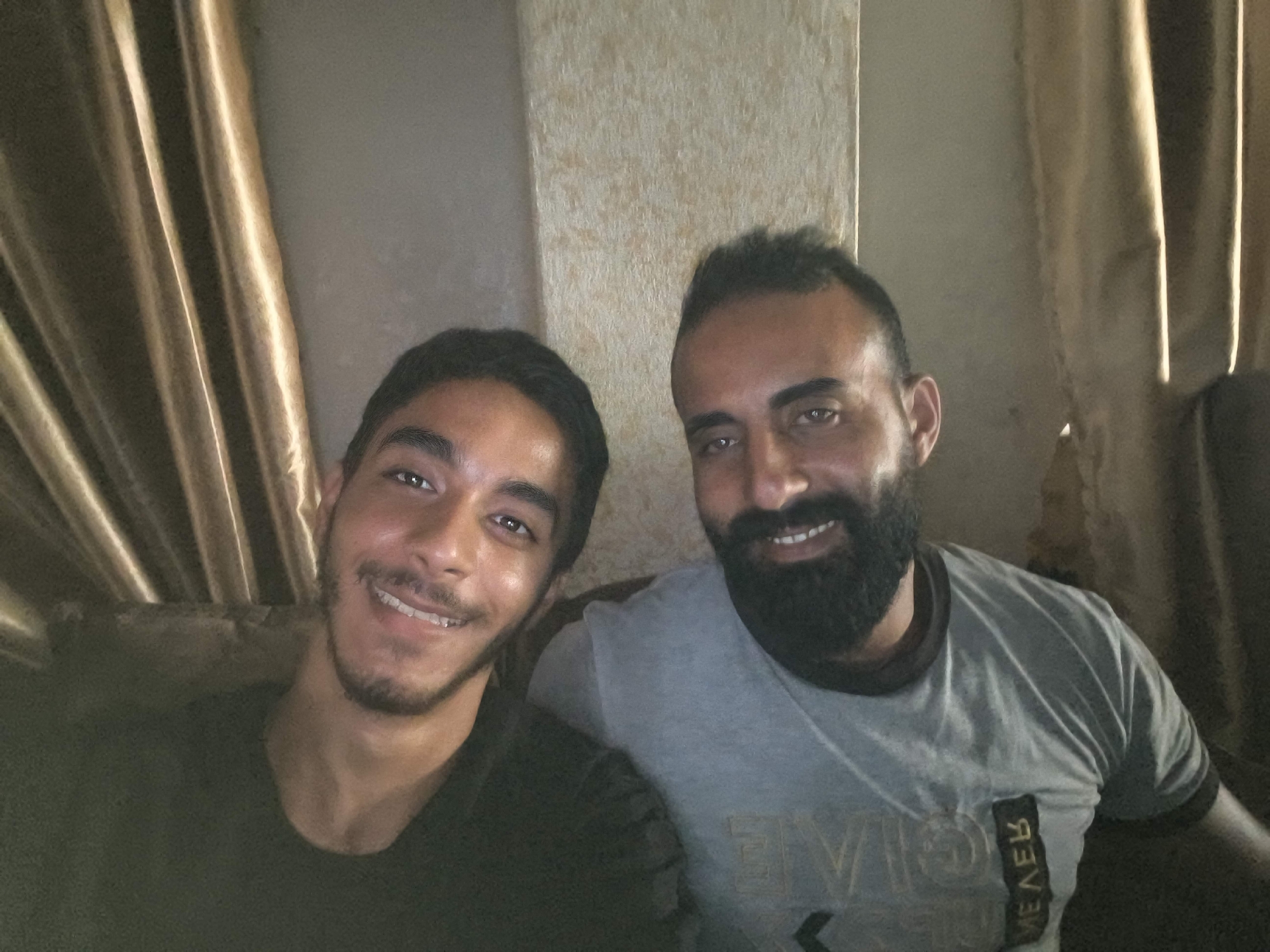 The image is a selfie taken indoors by two men. The man on the left, with dark hair and a thin beard, is smiling with his mouth open. The man on the right, with dark hair and a full beard, is also smiling. Both men are wearing t-shirts. The background consists of curtains and a textured wall. The lighting suggests an indoor environment, possibly a home. The men are positioned close together, indicating a friendly or familiar relationship. There are no other discernible objects or activities in the scene. The context of Gaza, Palestinian Territories, is external information and cannot be inferred from the image itself.