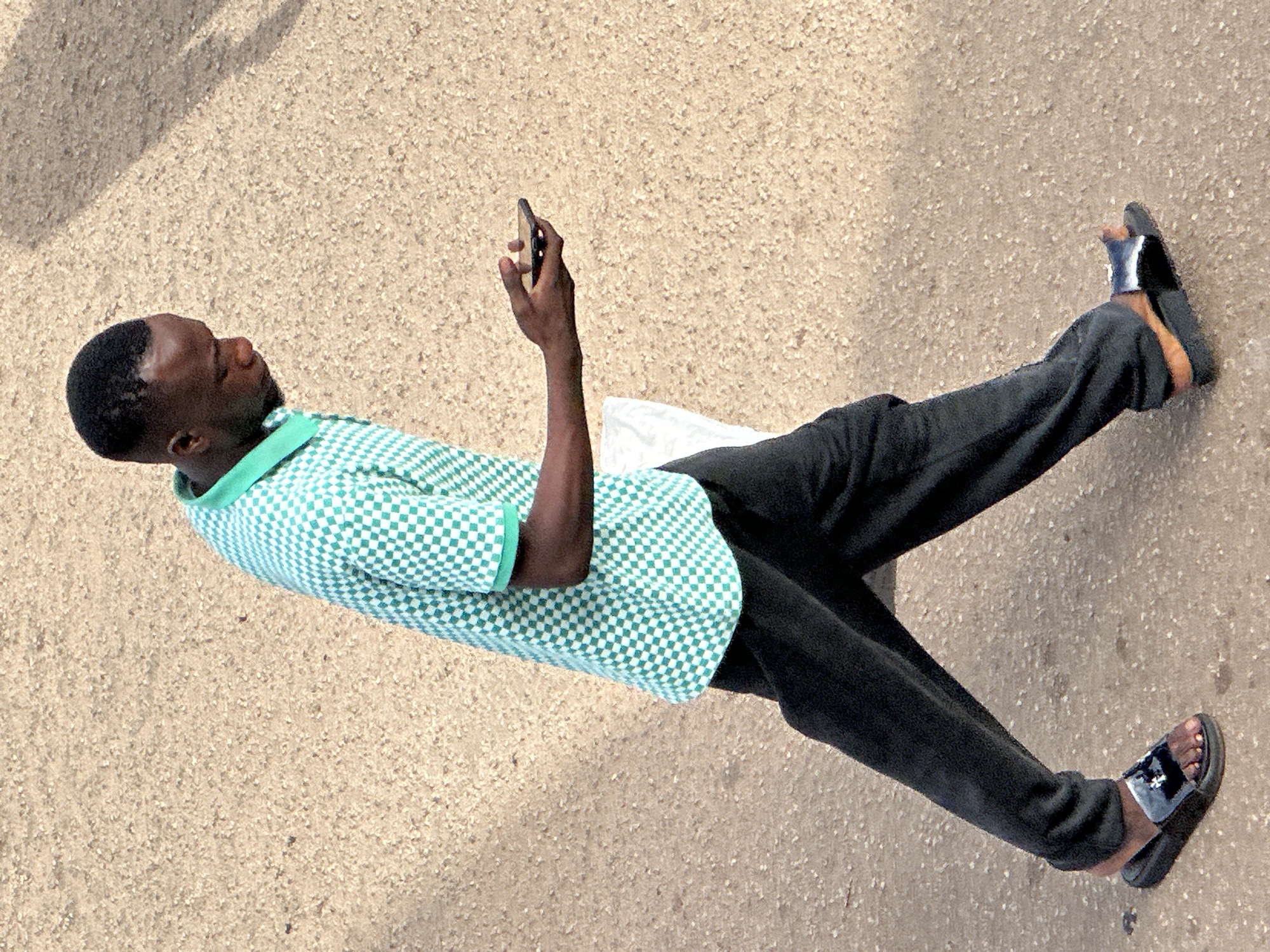 A man is walking on a dirt or gravel surface in Bukuru, Nigeria. He is wearing a teal and white checkered short-sleeved polo shirt and black pants. He has on black slide sandals with a silver embellishment. The man is holding a black smartphone in his right hand, looking down at it. A white bag is tucked under his left arm. The ground is a light brown color with small pebbles and shadows cast from an unseen source.