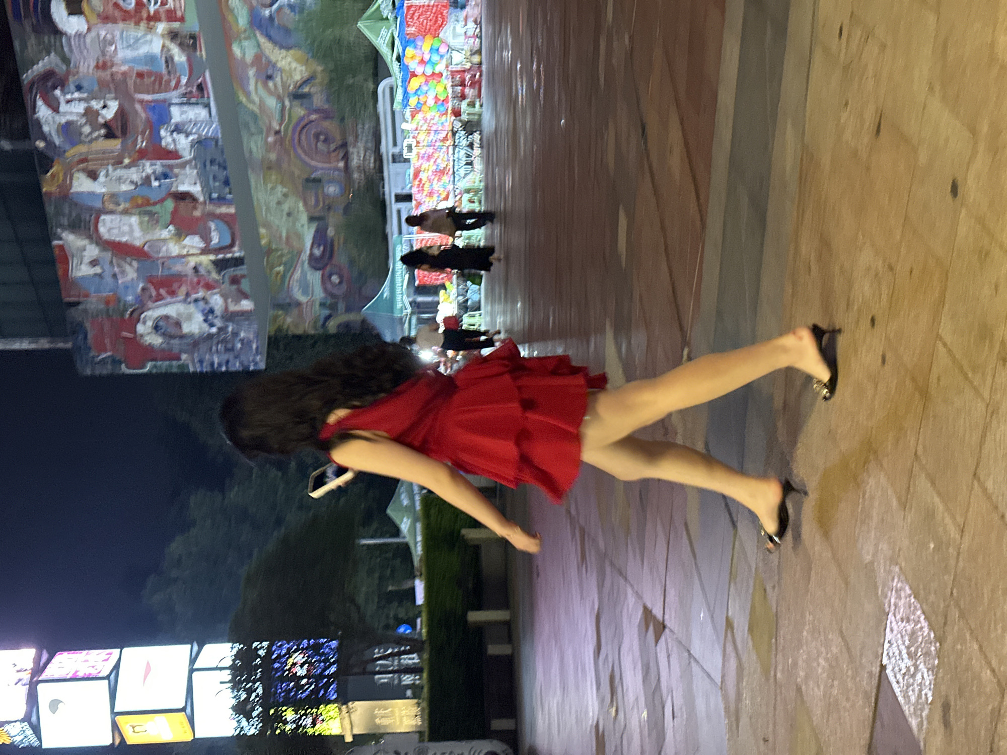 A young woman in a red dress walks on a wet paved area at night. She is looking at her phone. In the background, there are stalls with colorful balloons and other items, and a mural depicting abstract faces and shapes. The scene appears to be in a public plaza or street market in the Chongqing High-tech Zone, China.