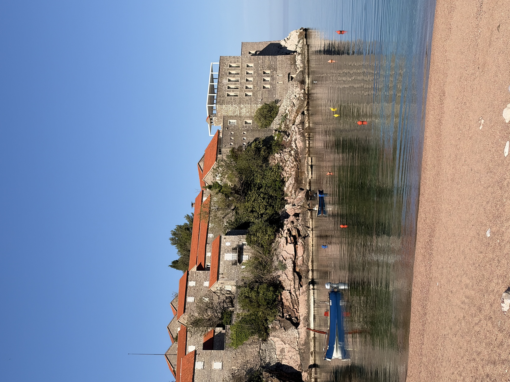 A serene coastal landscape, likely Sveti Stefan, Montenegro, is depicted under a bright, clear blue sky. On the left, a series of stone buildings with distinctive reddish-orange tiled roofs are built directly onto a rocky outcropping or island. These multi-story structures, characteristic of Mediterranean architecture, are interspersed with lush green trees and shrubs. One prominent building features a modern white rooftop terrace. Numerous windows, some with closed white shutters, are visible across the stone facades.
The buildings and their rocky foundations extend into calm, clear blue water that beautifully reflects the structures and the sky, creating ripple-distorted mirror images. Several small boats, including a blue and white vessel with an outboard motor, are moored in the water, along with a few orange and yellow buoys. To the right, a wide, fine-grained reddish-brown sandy beach curves along the shore.
The scene is bathed in natural daylight, suggesting late morning or early afternoon, with clear skies and no signs of inclement weather. The overall impression is one of tranquility and natural beauty, with no human activity or visible text to distract from the picturesque setting.
