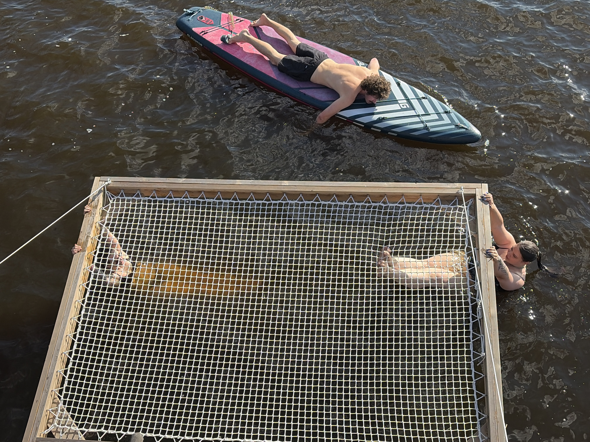 In Vidogoshchi, Russia, a high-angle shot captures a leisure scene on dark, rippling water. A wooden frame with a white net hammock extends over the water. Inside the hammock, two individuals are visible, one wearing a yellow garment and the other partially submerged. A third person, wearing dark shorts, lies on a paddleboard to the right of the hammock. The paddleboard is pink and gray with black stripes. The person on the paddleboard is supine, with their head facing the camera. Another individual is partially submerged in the water below the hammock frame, holding onto the edge with their hands. The water reflects sunlight, creating bright spots.