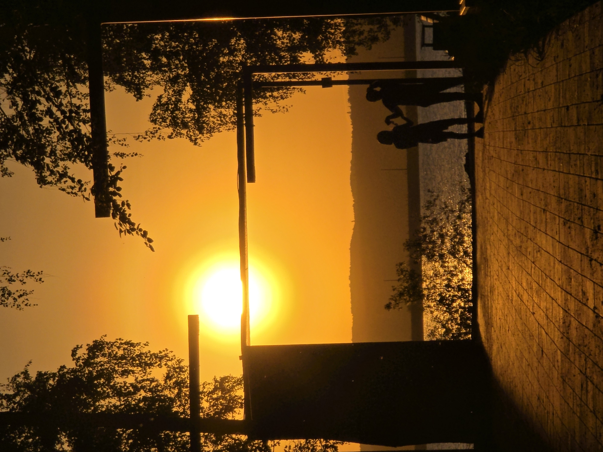 Captured from a low angle, the photograph depicts a vibrant sunset in Chaikovsky, Russia, reflected in a mirror-like surface. The scene includes silhouetted figures of an adult and a child. The environment appears to be an outdoor setting possibly a park or promenade, with trees flanking the sides. A brick pathway can be seen in the foreground. The dominant colors are shades of orange and yellow from the sunset, creating a warm, dramatic atmosphere.