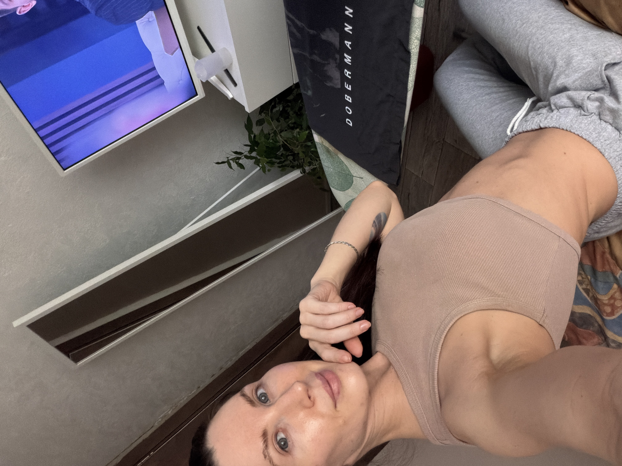 A woman with fair skin and blue eyes lies on a patterned surface, wearing a beige tank top and grey sweatpants. A black mat with "Dobermann" printed on it lies beside her. In the background, a television displays a blue screen with horizontal lines. A white cabinet stands to the side, next to a small green plant. A mirror on a grey wall reflects the scene. The image was taken indoors. There is no evidence to suggest the location is Perm, Russia.