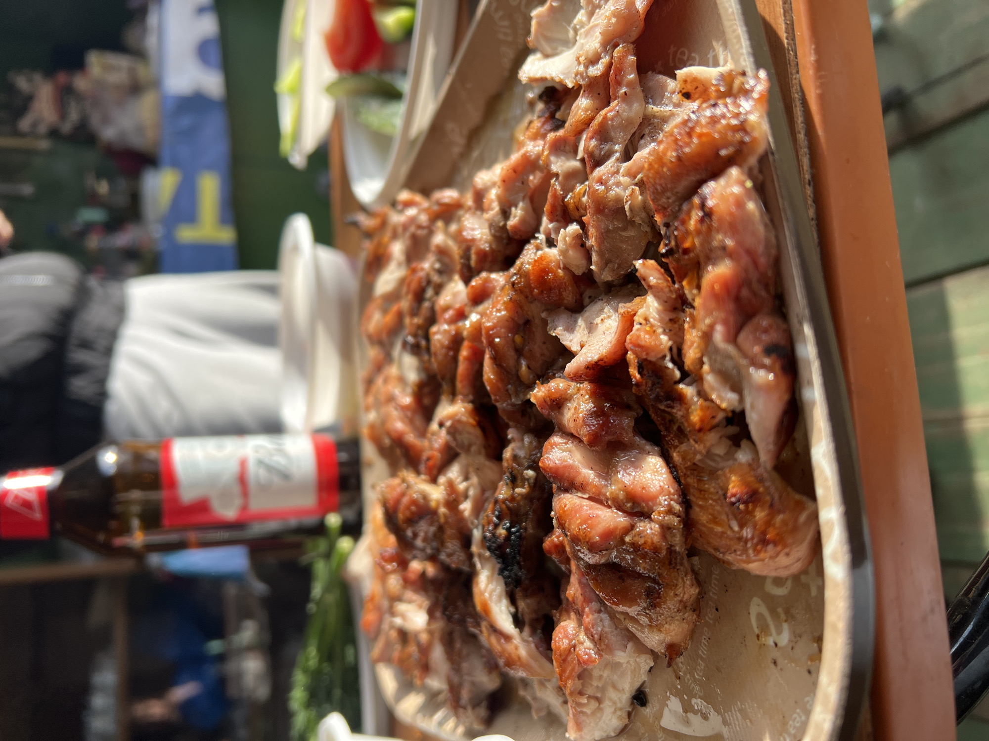 The image shows a close-up of a tray of grilled meat, likely chicken, with vegetables and a bottle of beer. The beer bottle is partially visible in the background, with a red label. The meat is piled on the tray in irregular pieces, appearing freshly cooked with a browned and slightly crispy exterior. The tray itself has a light brown color. The backdrop is blurred, showing a person wearing a dark jacket and a blue and white vertical sign. The environment appears to be outdoors, possibly at a food stall or event. The location, Noviy, Russia, is provided but not directly observable in the image.