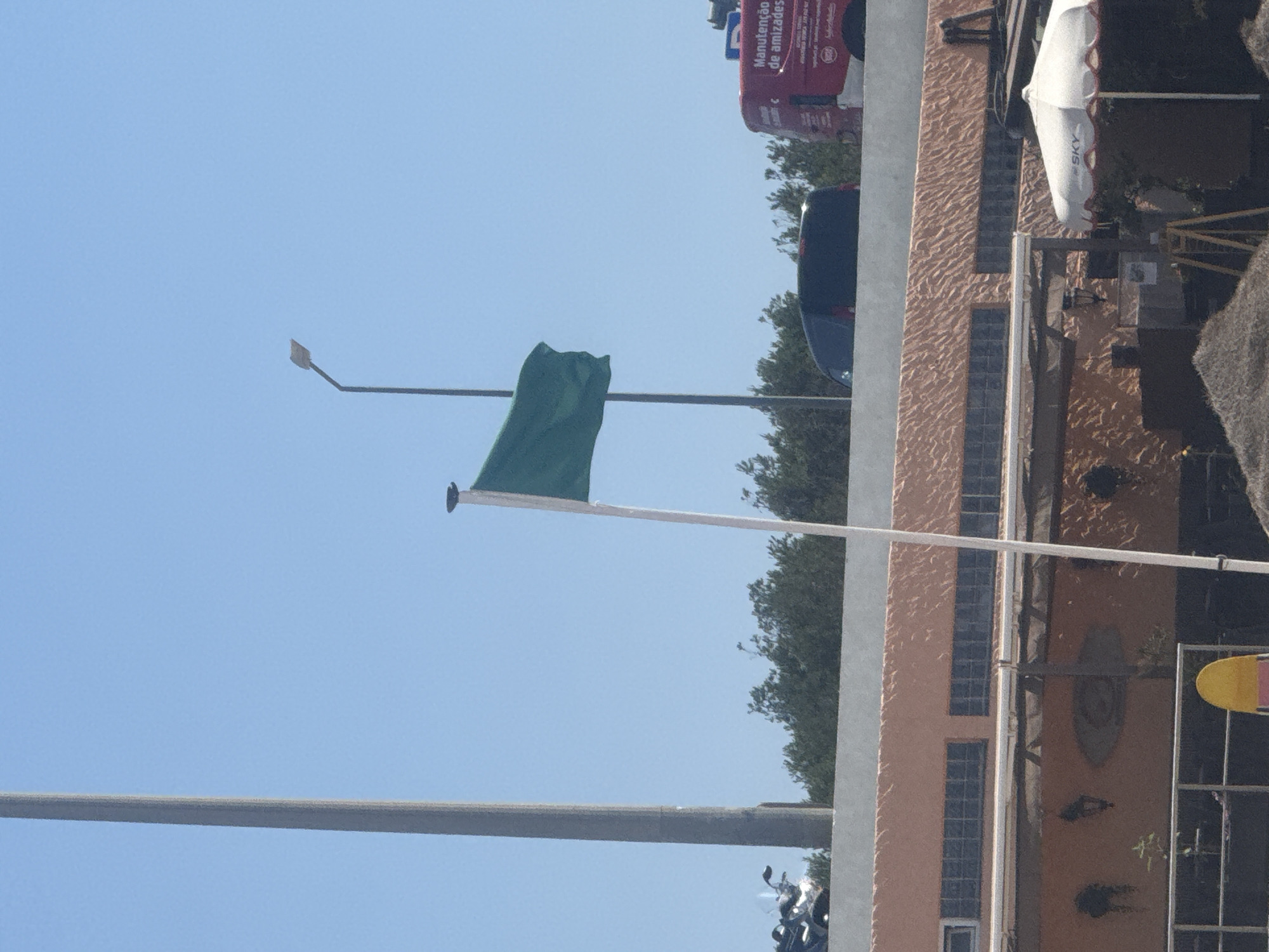 This image captures a bright, sunny day in Cascais, Portugal, likely near the coast. A large, tall, grey pole stands on the left side of the frame, partially obscuring the clear blue sky. In the middle ground, a green flag flutters on a white flagpole, indicating a safe condition, possibly for swimming or recreational activities. A streetlamp with an orange light is visible behind the flag. A dark-colored van is parked on an elevated road behind a low concrete barrier. To the right, a red vehicle with white text, which appears to be a maintenance or service truck, is also parked on the elevated road. The text on the red vehicle, partially visible, reads "Manutenção de amizades," suggesting it's related to maintenance services.
Below the elevated road, a peach-colored building with textured walls and rows of glass block windows is visible. Further down, parts of a beachside establishment are seen, including a white umbrella with "SKY" printed on it, a metal railing, and a yellow object, possibly a chair or a buoy. In the foreground, a pile of sand or thatched material suggests a beach or a shaded area. Trees line the horizon behind the building and the elevated road, creating a lush green backdrop against the sky. A motorcycle is partially visible on the left, parked near the grey pole. The overall scene depicts a public space, possibly a promenade or a beach access area, on a clear day.