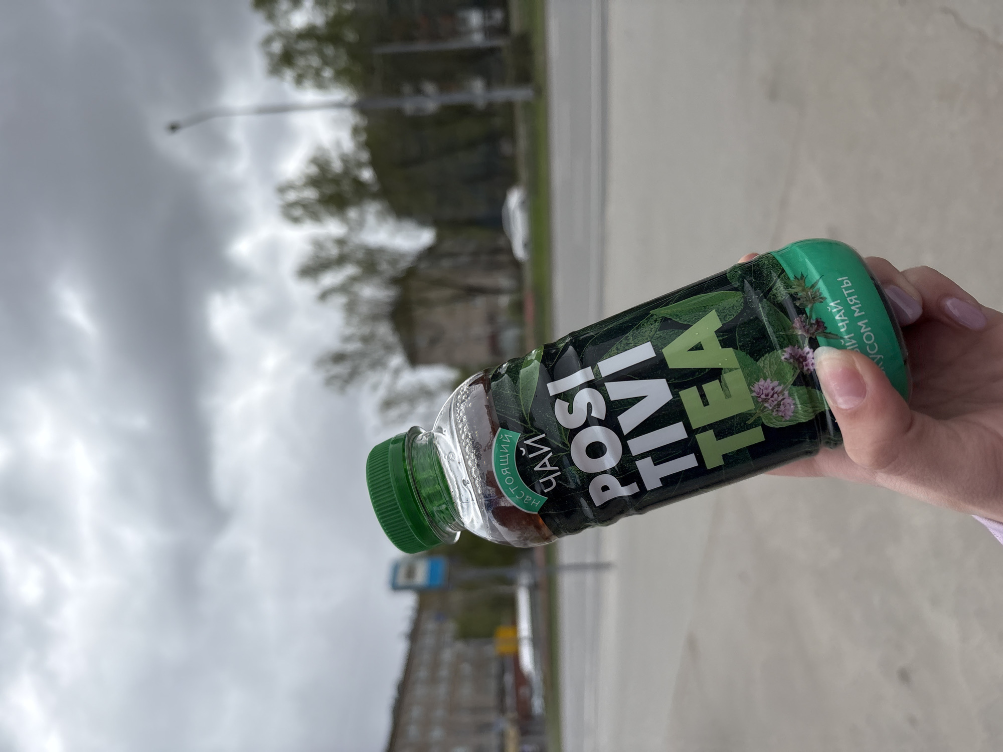 The image shows a hand holding a bottle of "Positiv Tea" against a cloudy sky and a blurred urban background. The bottle is green and black with white lettering. The background suggests an urban setting, potentially Perm, Russia, given the Cyrillic text on the bottle. The area appears to be a street with buildings and trees visible in the distance. The weather is overcast.