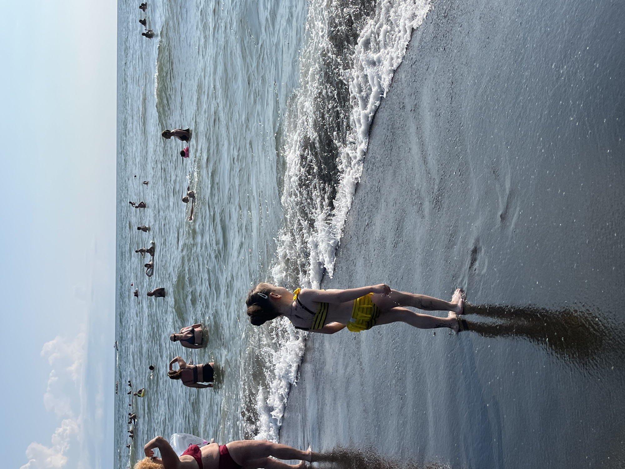 This is a vertical, eye-level, medium shot of a beach with many people in the water and on the shore. The scene is set in Ureki, Georgia, under a bright, clear sky, suggesting it's a sunny day. The water is a deep blue-green and choppy with waves breaking on the shore, creating white foam. The sand appears dark and wet near the water's edge.
Numerous individuals are visible, most of them partially submerged in the water. Some are swimming, while others appear to be wading or standing. In the foreground, a young person wearing a yellow and black striped bikini and a black life vest is lying on their side near the water's edge, facing away from the camera. Their legs are extended towards the water, and they appear to be relaxed. Further down the shore, another person in a red bikini is lying on their back with their legs extended toward the water, a pool float visible beside them. In the distance, many other people are scattered throughout the water, some of whom are also in bathing suits. The overall mood seems to be one of leisure and recreation on a summer day at the beach. No text is visible in the image.