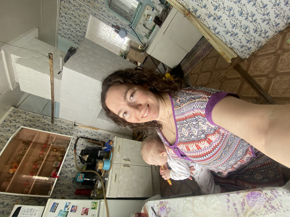 A woman holds a baby in what appears to be a kitchen or utility room in Uzyar, Russia. The woman, with brown curly hair, smiles directly at the camera while holding the baby in her left arm. She wears a multi-patterned top with purple trim. The baby, visible from the back and left profile, wears a white long-sleeved outfit and holds a yellow object in its right hand, appearing to be chewing on it. The baby has light-colored hair.
The room features blue floral patterned wallpaper. To the immediate left of the woman and baby is a white table with a light-colored tablecloth adorned with purple floral designs, upon which a glass with a reddish liquid is visible. Further left, a white refrigerator with various magnets and notes is visible. Above a light-colored base cabinet with two doors and a counter, a wall-mounted glass display cabinet holds multiple decorative items such as small bottles and teacups. The counter below is cluttered with jars, bottles, a black plastic bag, and a blue container.
In the background, a large, multi-tiered white structure, consistent with a traditional Russian stove, occupies a significant portion of the room, extending from floor to ceiling. A wooden beam is suspended horizontally from this structure. To the right of the stove, an older-style sink unit with a light-colored base cabinet and a decorative mirror or panel above it is visible. A window with a light-colored curtain is present in the background. The floor is covered with brown diamond-patterned tiles, with a patterned rug partially visible on the right.