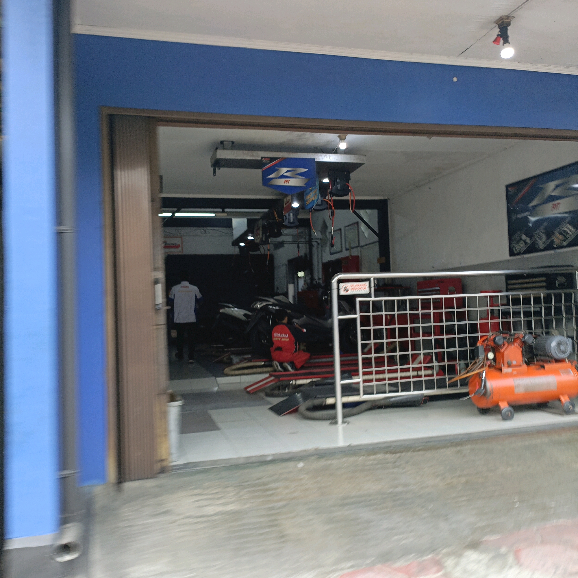 The image captures a view from the exterior of a motorcycle repair shop, likely located in Salatiga, Indonesia, given the context of similar imagery. In the foreground, a blue pillar and a brown ribbed sliding door are visible on the left and center respectively. A metallic fence with a white frame encloses an area to the right. Inside this enclosure, an orange air compressor with a black motor sits on a grey platform.
Through the open doorway, the interior of the repair shop is partially visible. The shop appears to be a well-lit space with a white tiled floor. A person wearing a white shirt and dark pants stands near the entrance, facing away from the camera. Further inside, another person in a red uniform with the "YAMAHA" logo is kneeling next to a motorcycle. Several other motorcycles and various tools and equipment are visible in the background, including red tool chests. Above the work area, a blue sign with a white "R" logo and the word "PIT" is suspended from the ceiling, along with lighting fixtures. A poster with a motorcycle design is mounted on the wall in the upper right portion of the interior. The overall impression is of an active motorcycle service center.
