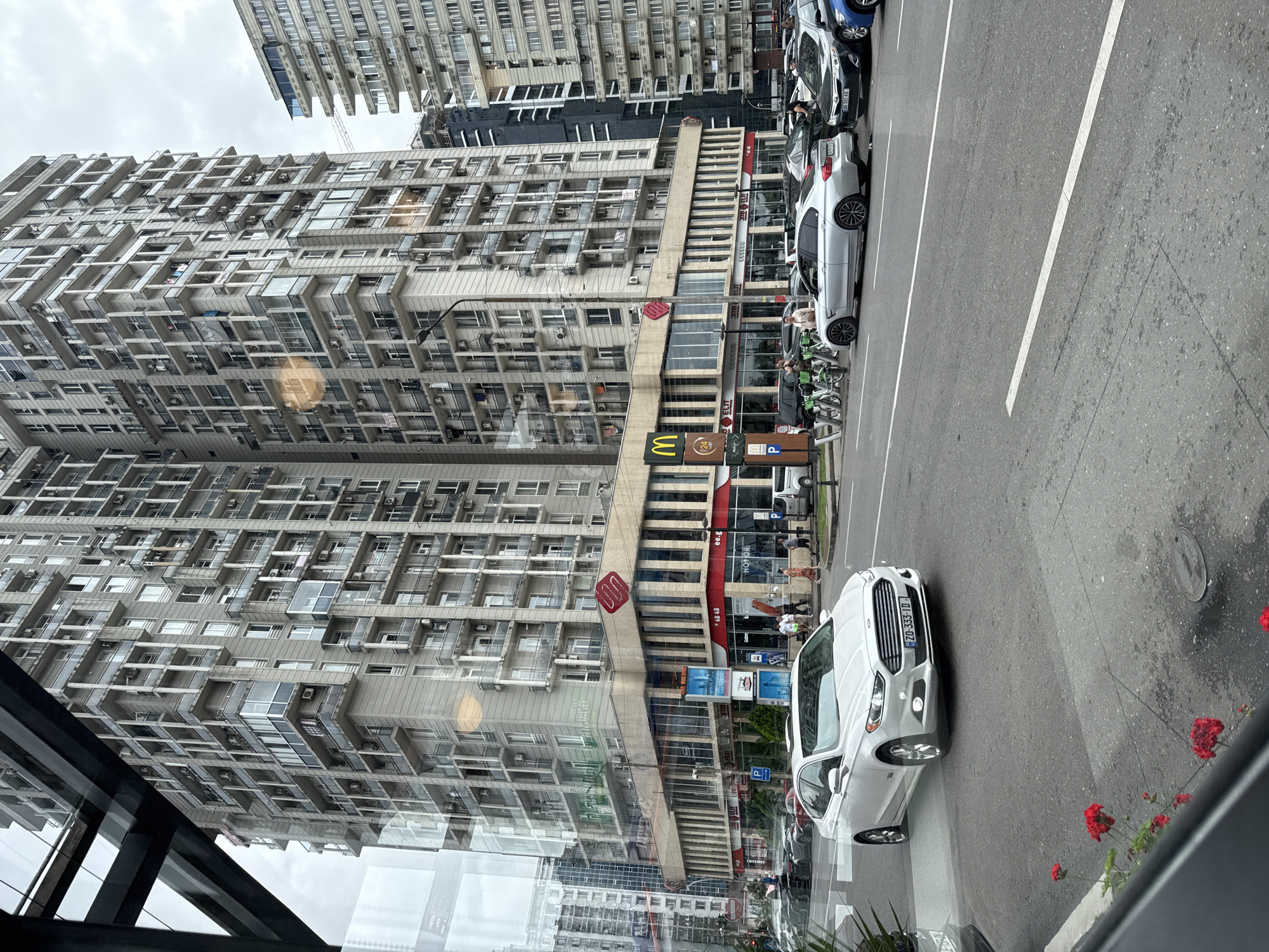 The image captures a street scene in Batumi, Georgia, dominated by a towering, multi-story apartment building with numerous balconies and windows. A McDonald's restaurant with its signature golden arches is visible on the ground floor of this building, alongside other businesses. Several cars are parked and driving on the street, including a white Ford sedan in the foreground. Bicycles are also lined up along the sidewalk. The weather appears overcast. The reflection of the sky and parts of the environment are visible on the glass from which the photo is taken, suggesting the vantage point is from inside a building or vehicle looking out. Red flowers are blooming in the lower left corner of the frame.