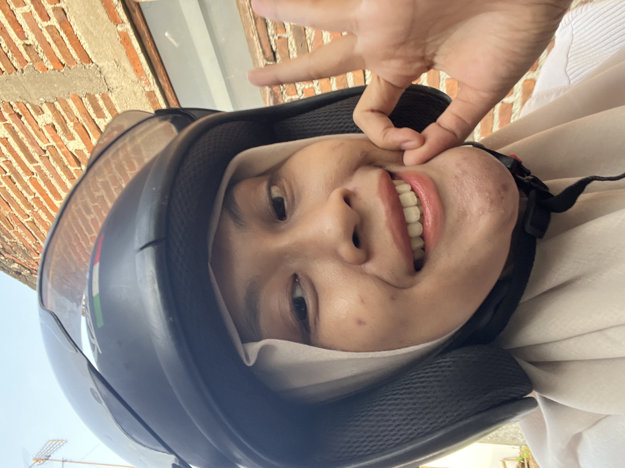 A woman wearing a black helmet and a light-colored hijab is taking a selfie outdoors. She is smiling, showing her teeth, and making an "okay" gesture with her right hand. She has some acne on her chin. Behind her, a brick wall is visible. The scene appears to be in a residential area, possibly in Kecamatan Pati, Indonesia, given the style of the brickwork and the presence of what looks like an antenna in the background.