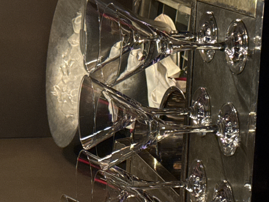 Three martini glasses sit on a metallic surface. Ice is visible in the background, with a white towel draped nearby. The glasses are arranged in a row, angled upward and facing the same direction. Their stems are reflective, adding to the overall sheen. The surface appears to be part of a bar or a display case.