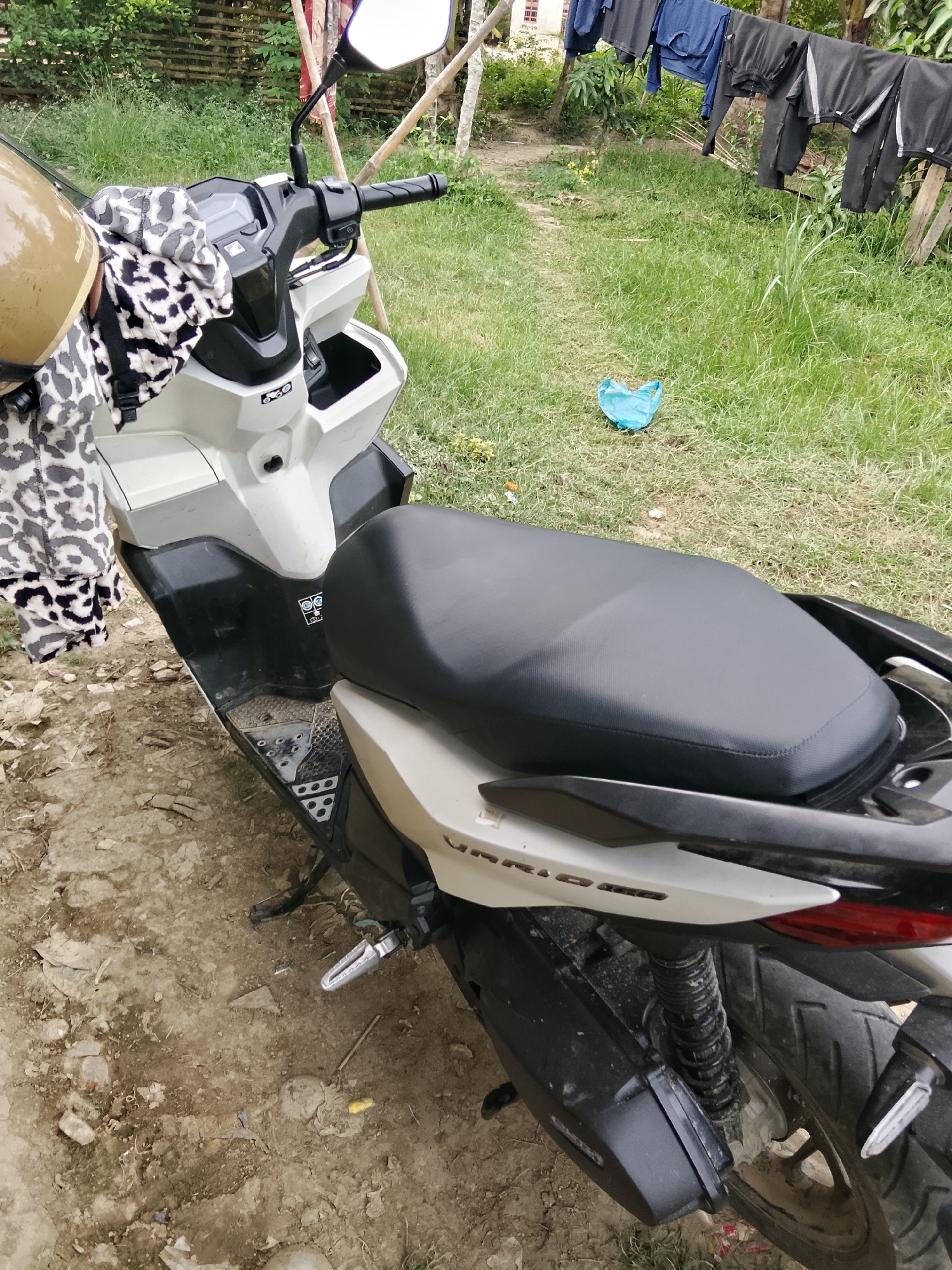 This is a medium-angle, eye-level, exterior shot of a white and black scooter parked on a dirt and gravel pathway. The scooter is the primary subject, facing slightly to the right. A tan helmet with a black and white leopard print cloth draped over it rests on the handlebars. Behind the scooter, a clothesline with dark colored garments is strung between wooden poles. The background consists of green grass, some trees, and a dirt path. The scene appears to be in a rural or semi-rural area, possibly in Kruenglingka Barat, Indonesia, based on the provided context.
