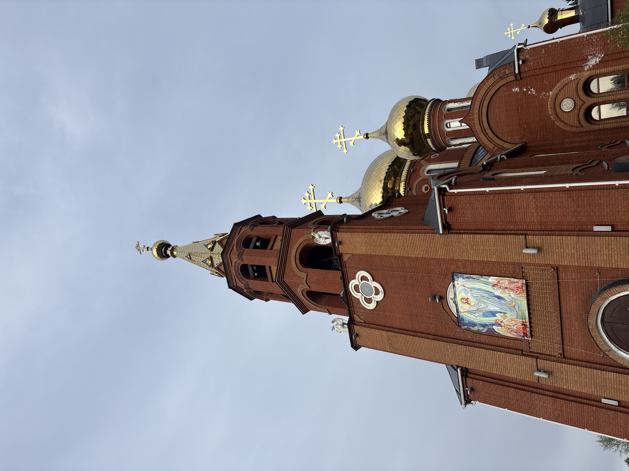The image shows the exterior of the Church of the Holy Great Martyr George the Victorious in Chaikovsky, Perm Krai, Russia. The church is made of red brick, with golden domes topped with crosses. A tall tower with a spire rises to the left, also adorned with a cross at its peak. An image of what appears to be Jesus with angels hangs on the brick facade. The sky is overcast.